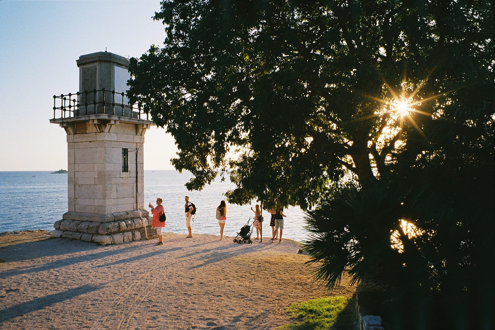 A lighthouse on a coastal path with people walking and enjoying the view, as the sun shines through the trees.