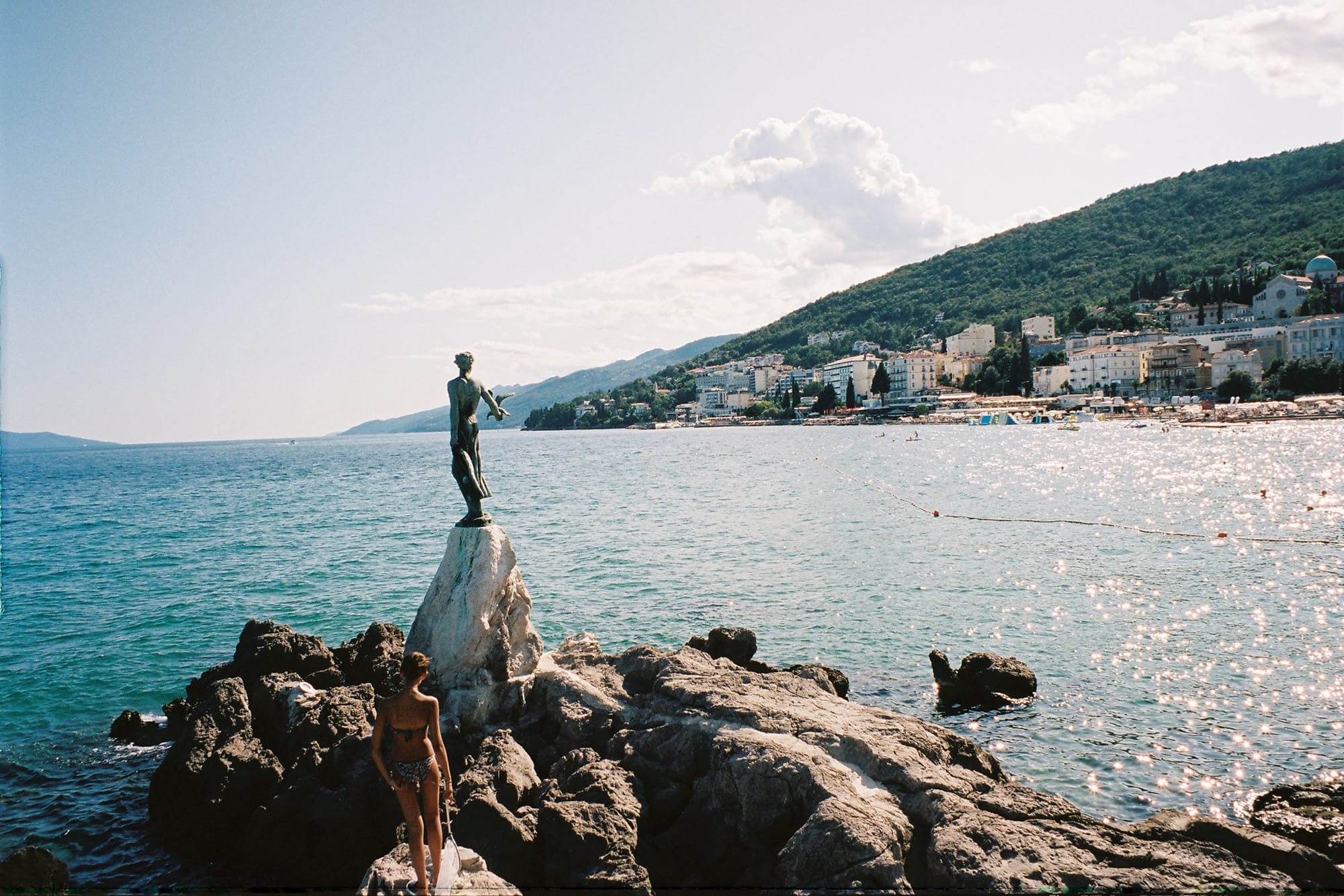 A statue and landmark on a rocky outcrop by the sea, with idyllic Opatija in the background.