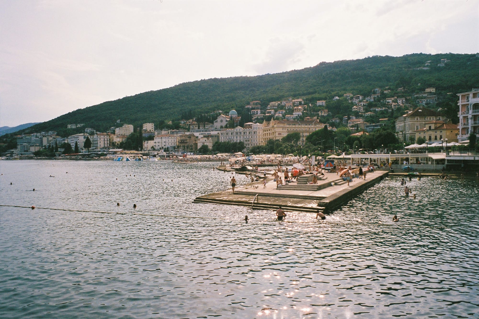 A pier extending into the sea with people swimming and sunbathing, and a coastal town with buildings in the background.