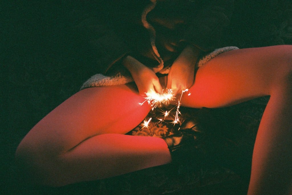 Young woman sitting provocatively in a dark setting, with a sparkler in hand.