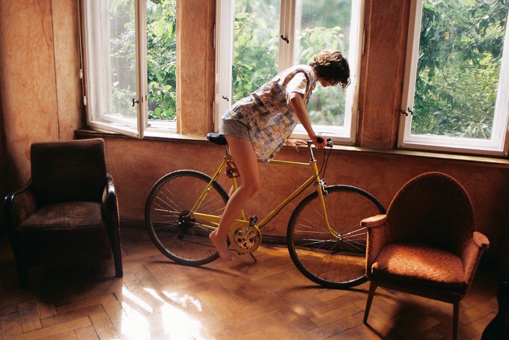 Young woman in a patterned button-up, sitting on a yellow bicycle indoors, in front of a row of windows.