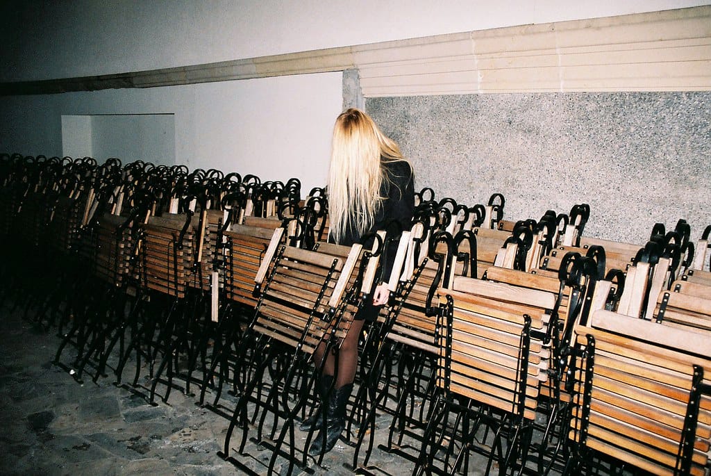 Young woman with long blonde hair standing in a room filled with rows of wooden benches.