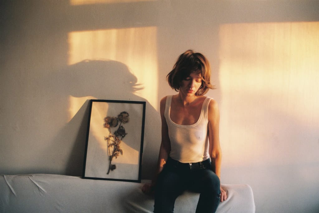 A young woman, sun-kissed, sitting on a white sofa, in a white bodysuit and black jeans, next to a framed dried flower art piece.