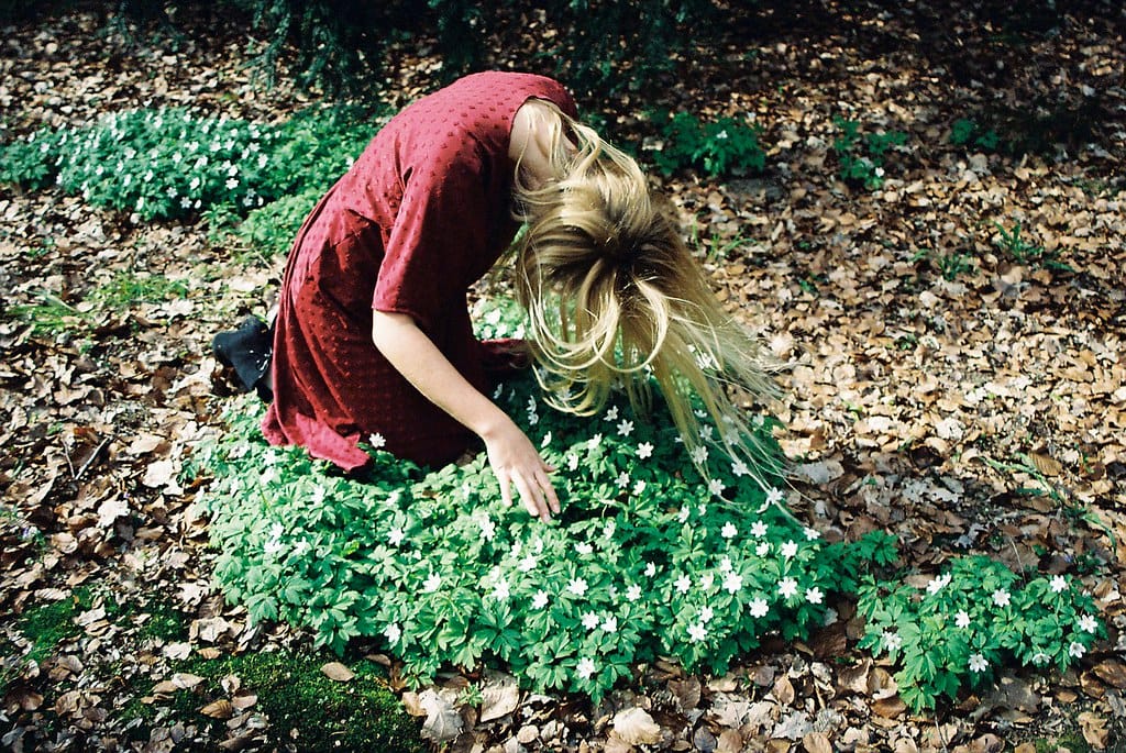 Blonde in a red dress bending down to touch white flowers in a forest.