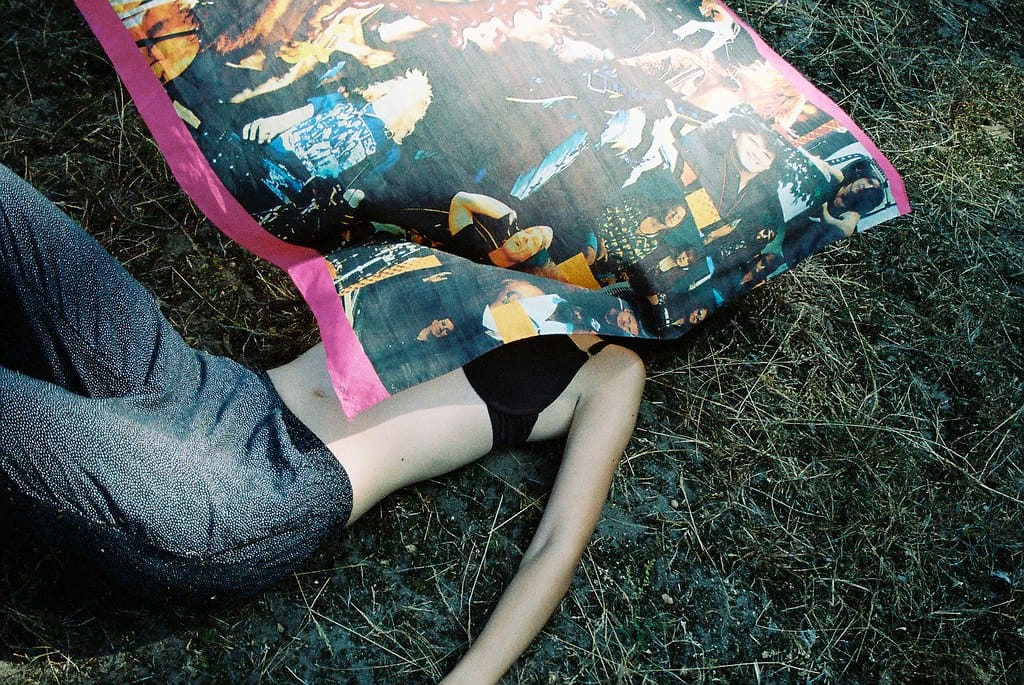 Young woman in a bra lying on grass as someone waves a colorful blanket with pink trim over her.