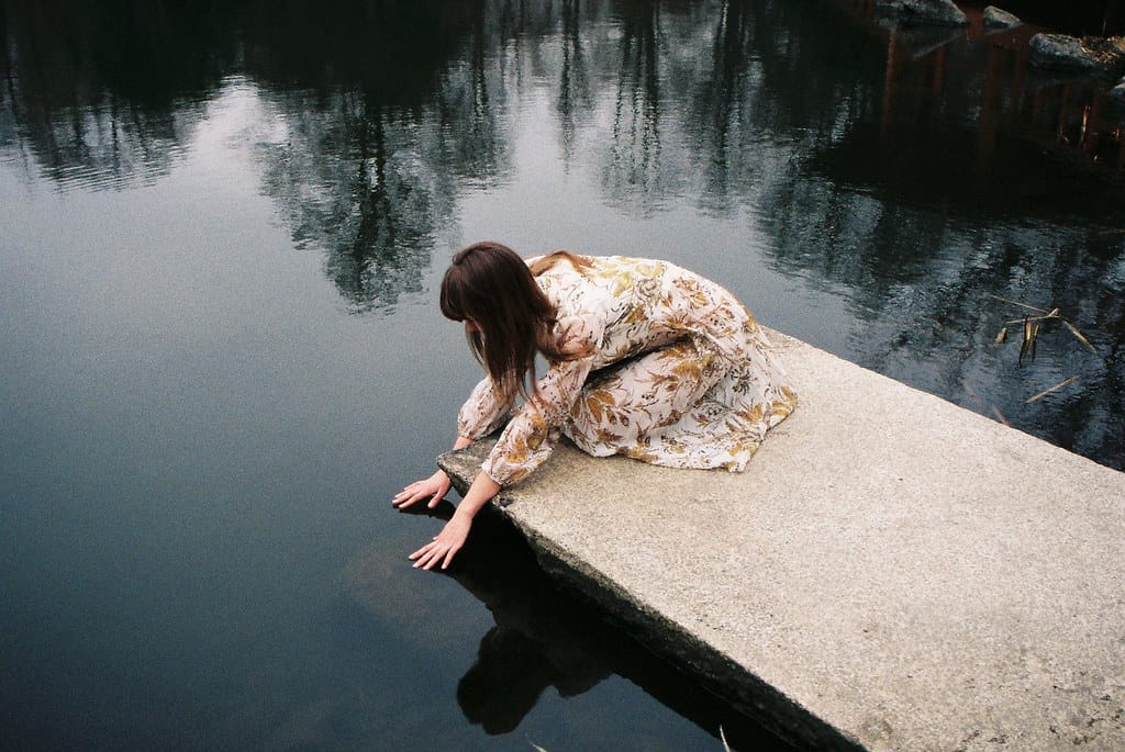Brunette in a floral dress reaching out over a still pond.