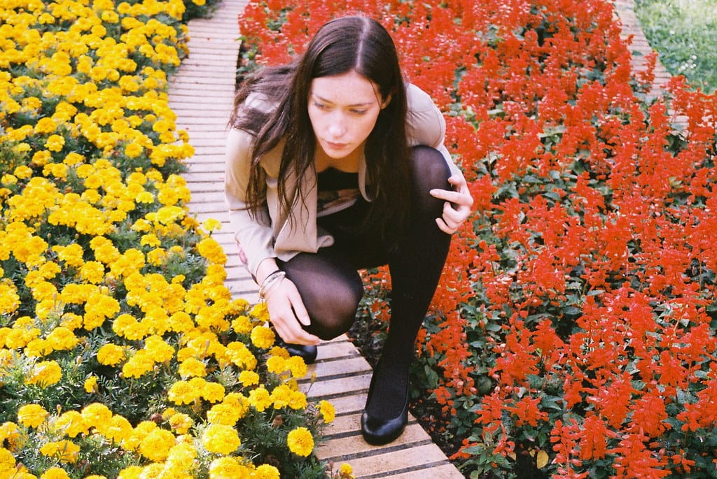 Young woman with long dark hair squatting on a wooden walkway amidst vibrant yellow and red blooms.