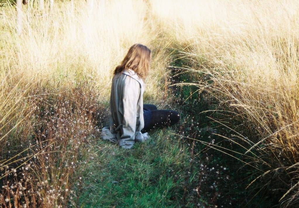 Woman with long hair sitting in a natural area, surrounded by tall, golden blades of grass.