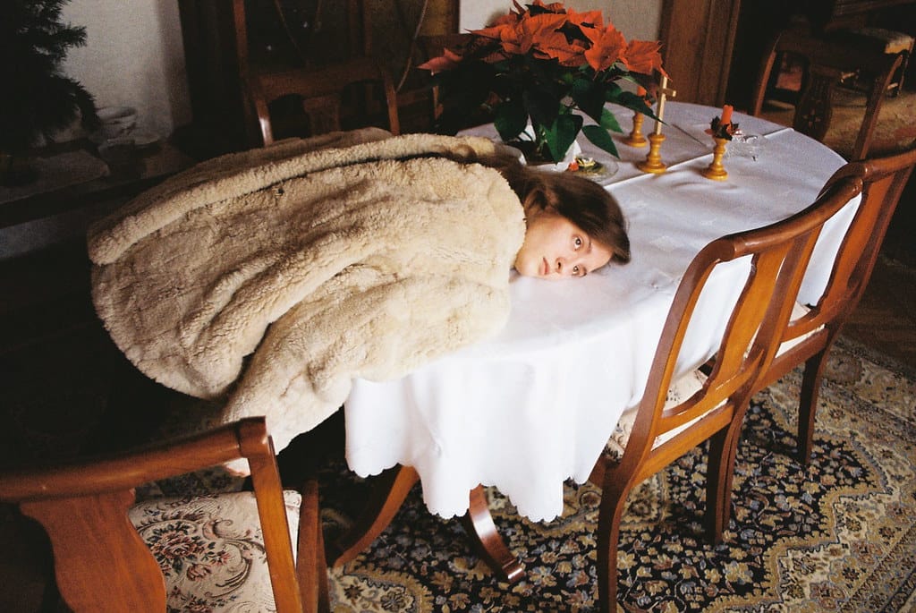 Girl in a beige fur jacket sitting on a chair with her upper body extended on a table, face resting on the side.