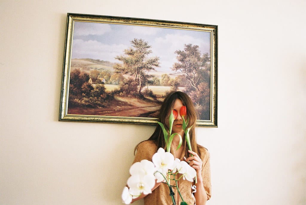 Young woman stands in front of a landscape painting, covering her eyes with flowers.