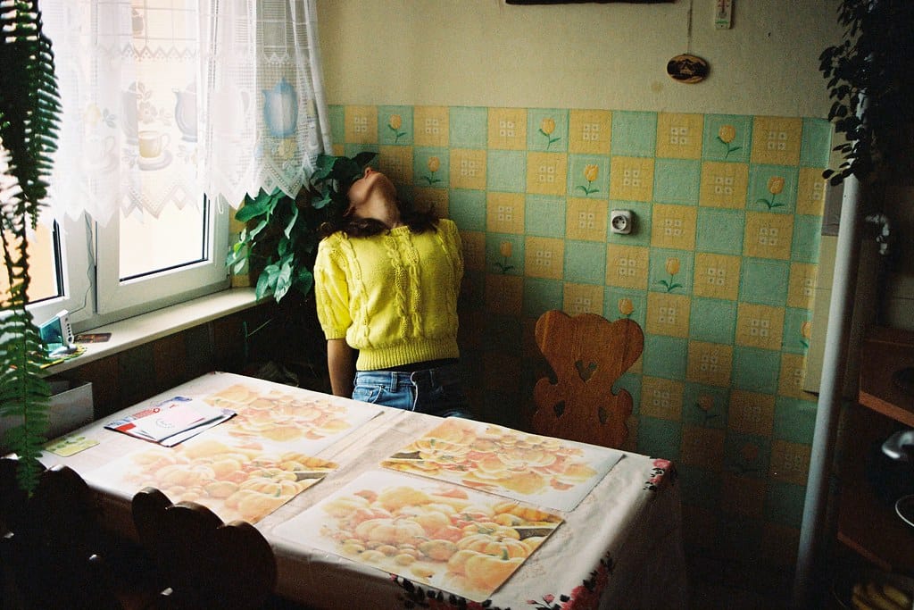 Young woman in a yellow sweater leaning into the corner of a kitchen with floral wallpaper.