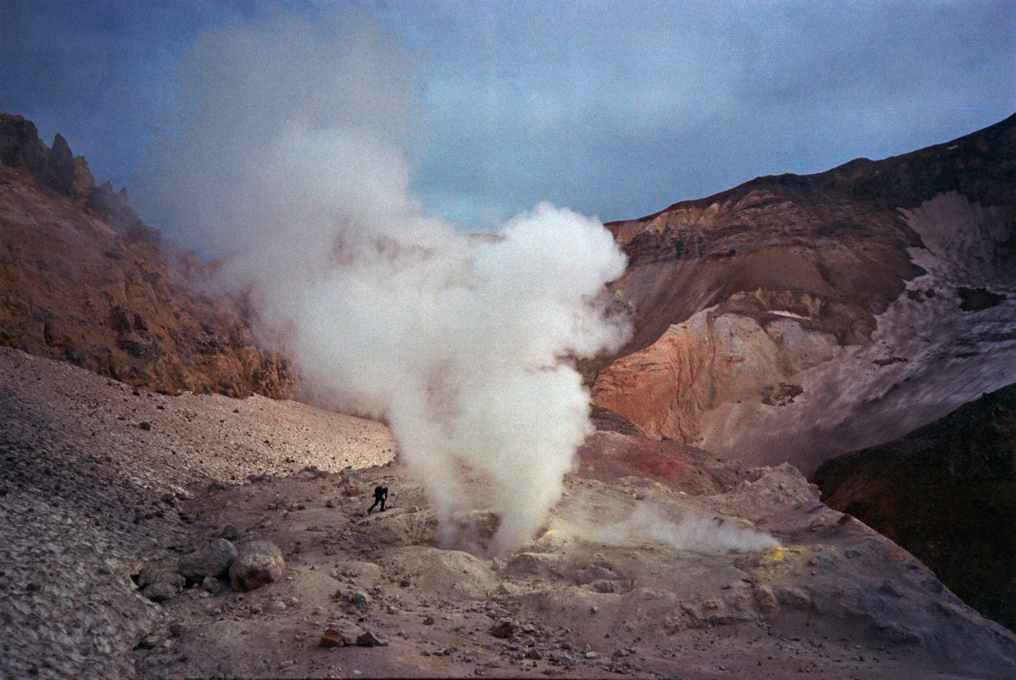 A person hiking near a large geyser emitting steam in a volcanic crater with rugged, rocky terrain and misty mountains in the background.