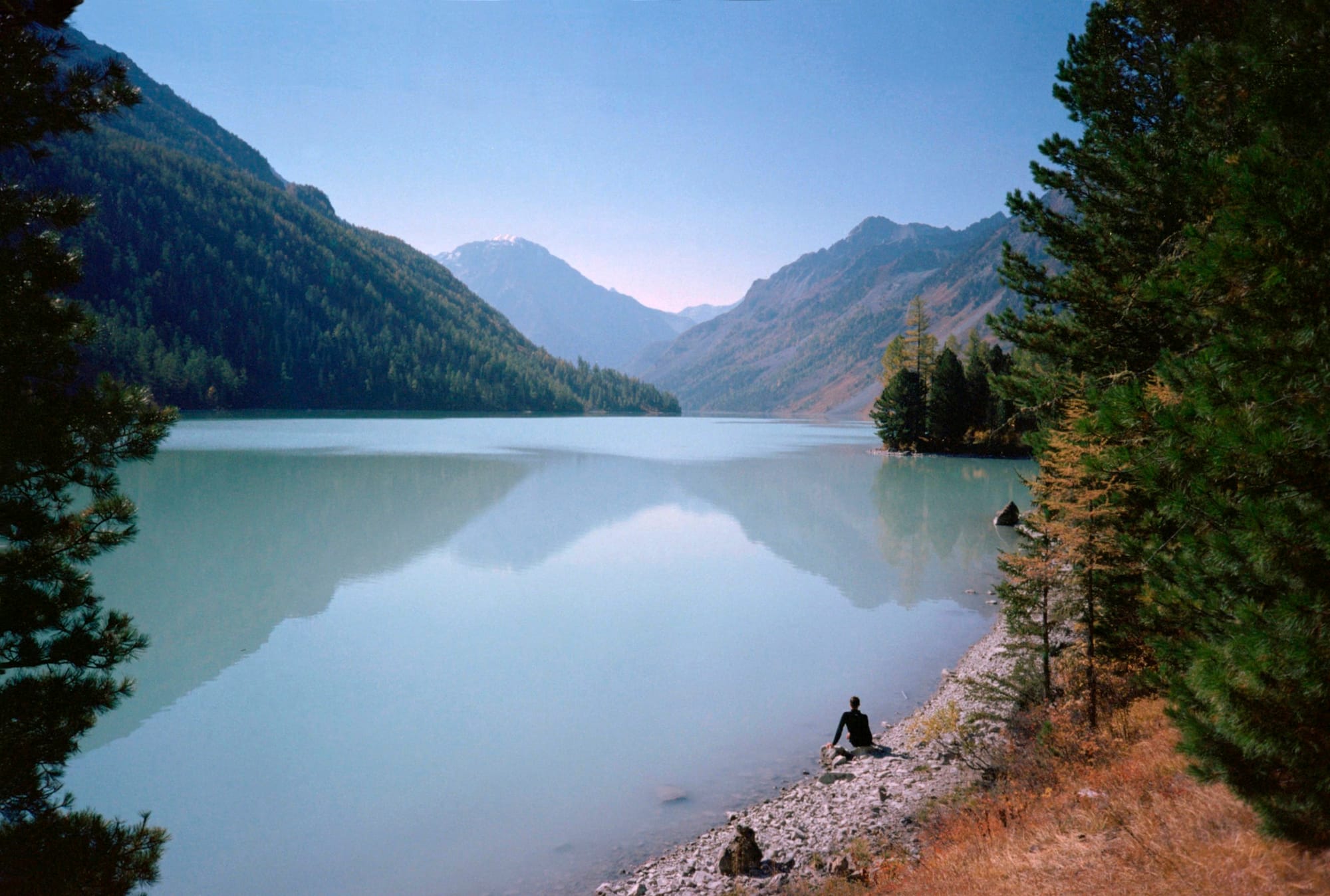 A person sitting on a rocky shore by a serene, clear lake surrounded by evergreen trees and mountainous terrain under a clear sky.
