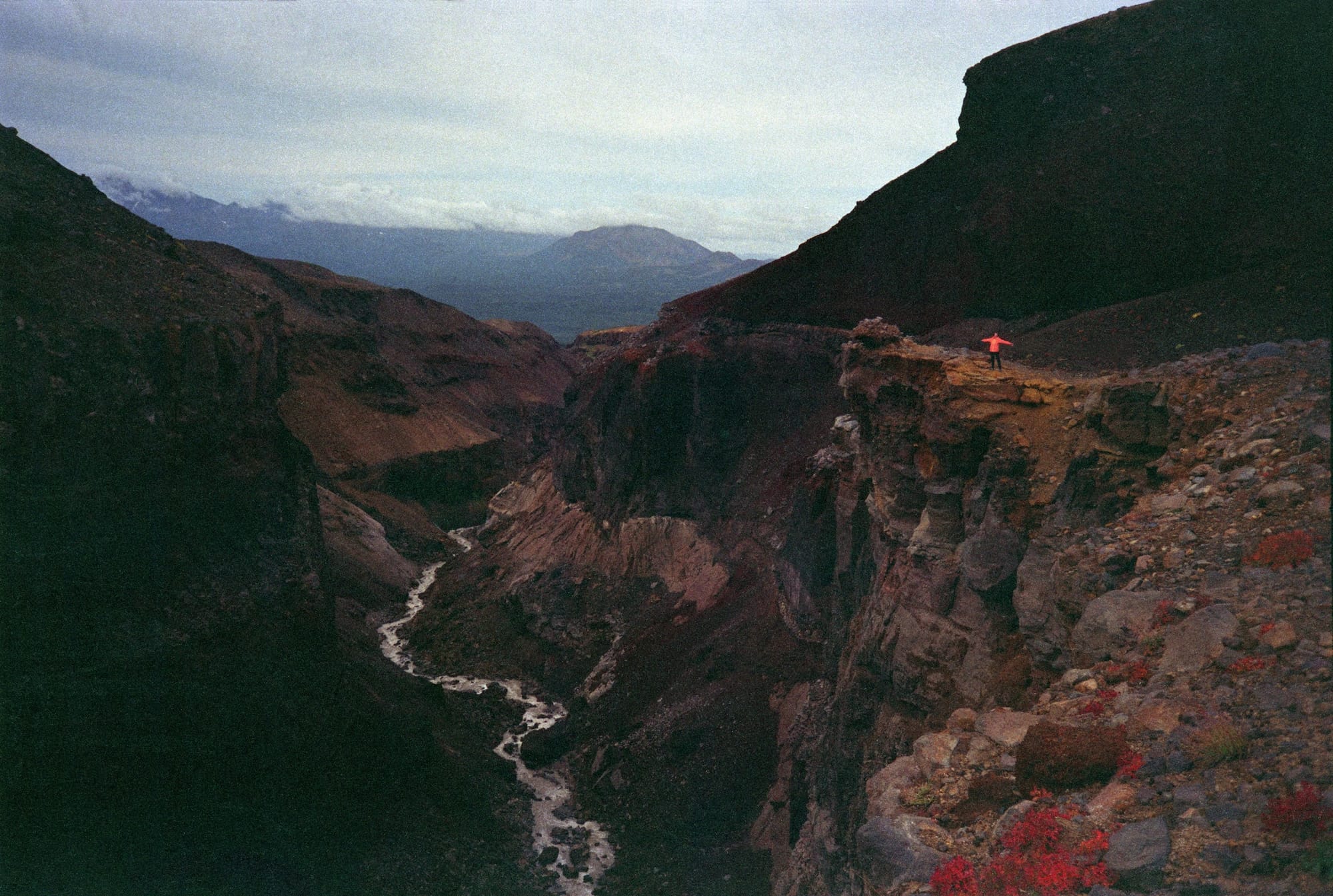 A person standing, arms spread, on a rocky ledge overlooking a deep, rugged canyon with a winding river below and distant mountains under a cloudy sky.