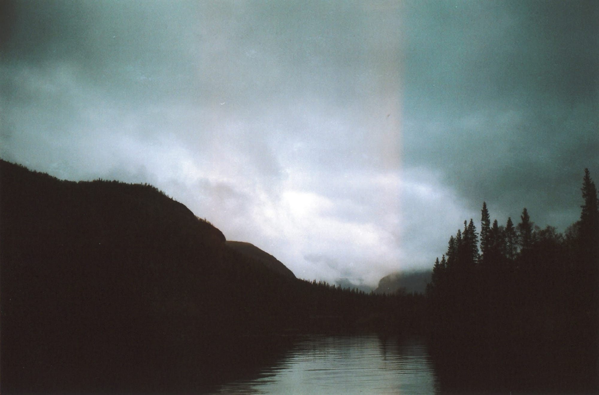 A serene lake reflecting a cloudy sky and misty mountains, surrounded by dark, silhouetted evergreen trees.
