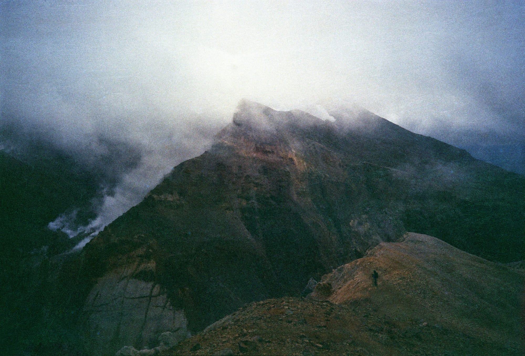 A lone figure standing on a rocky hillside at dusk, with misty mountains in the background under a dimly lit sky.