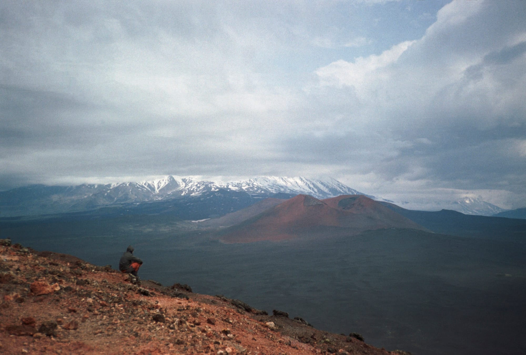A person sitting on a rocky hillside overlooking a vast, cloudy landscape with distant snow-capped mountains under a dramatic sky.