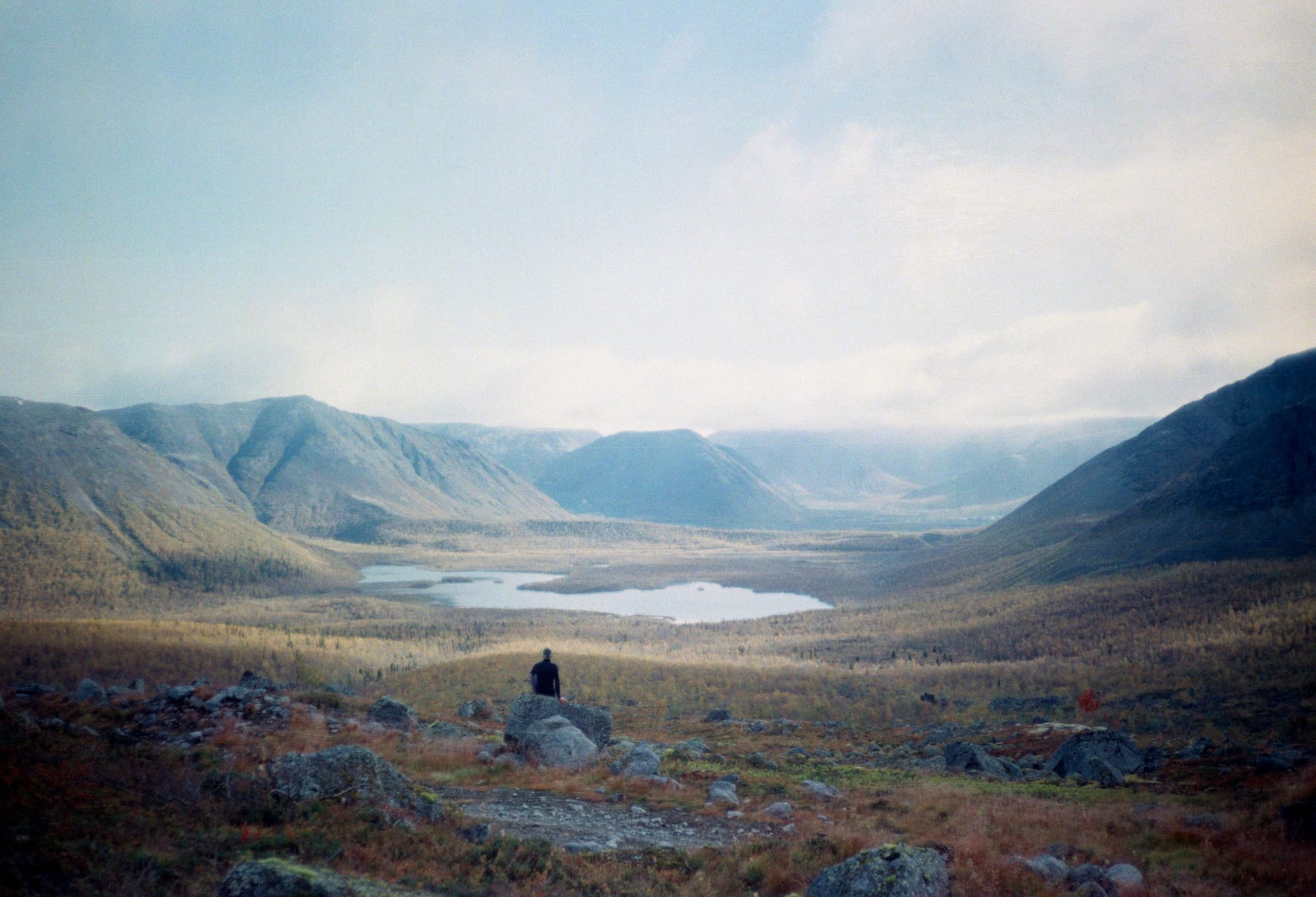 A person sits on a rock overlooking a vast, open valley with a small lake and rolling hills under a cloudy sky.