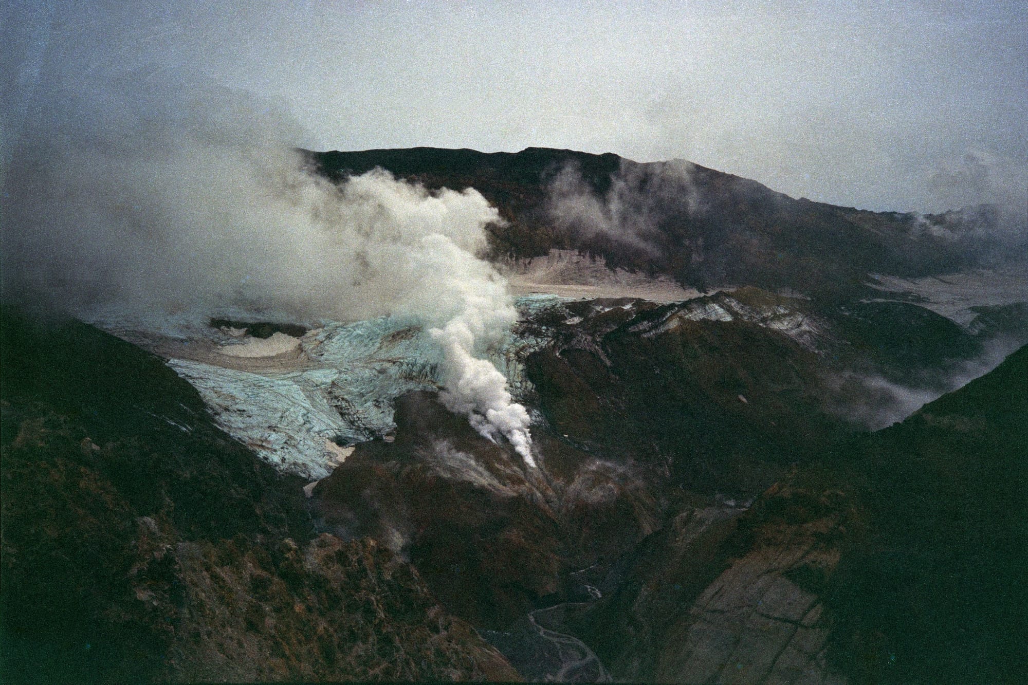 Aerial view of a volcanic crater emitting steam, with rugged, barren terrain.
