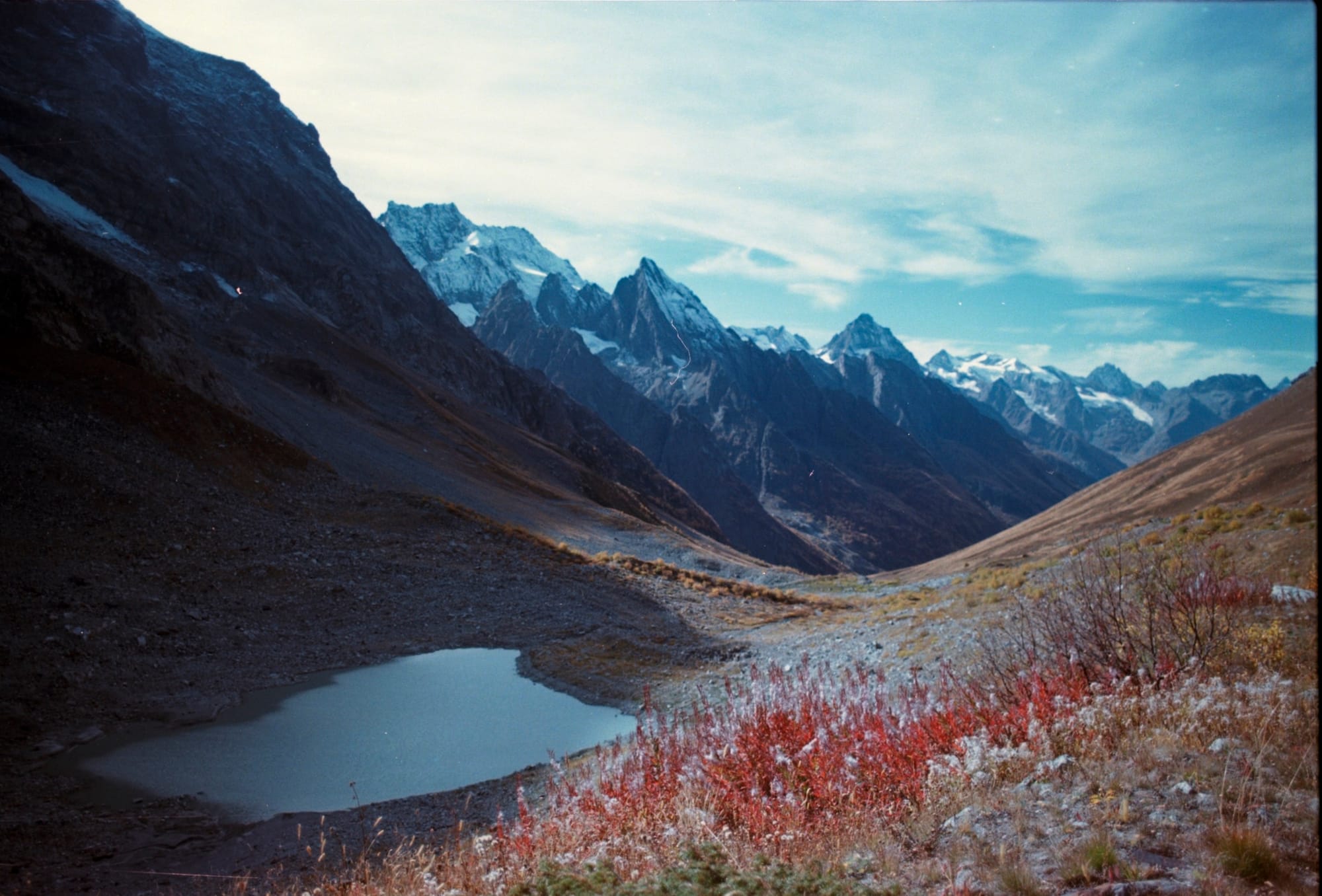 A serene mountain landscape with rugged peaks, a small alpine lake, and sparse vegetation in the foreground under a partly cloudy sky.