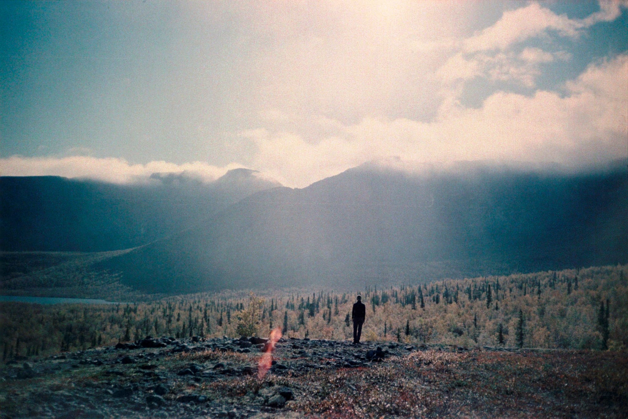 A person standing on a rocky hillside overlooking a vast, misty valley with distant mountains under a partly cloudy sky.