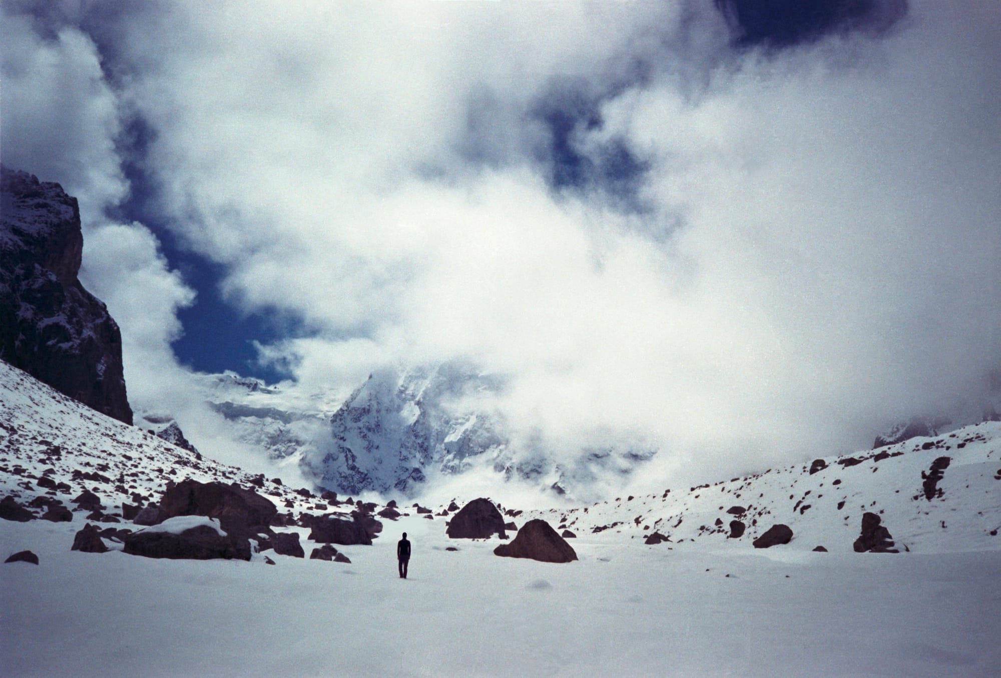 A lone person standing on a snow-covered landscape with misty, rugged mountains in the background under a cloudy sky.