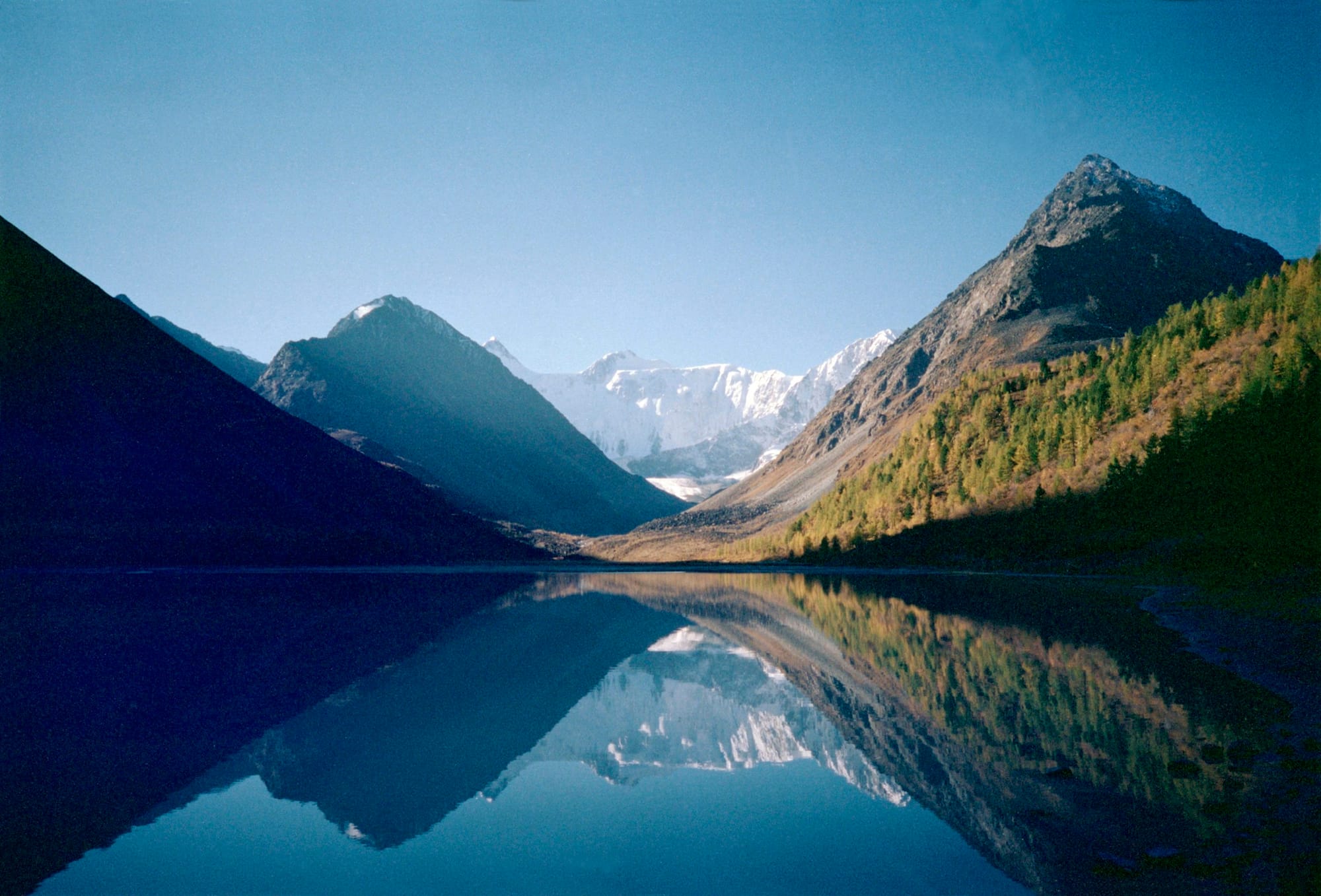 A serene mountain landscape with snow-capped peaks reflected in a calm, dark blue lake, surrounded by evergreen trees under a clear sky.