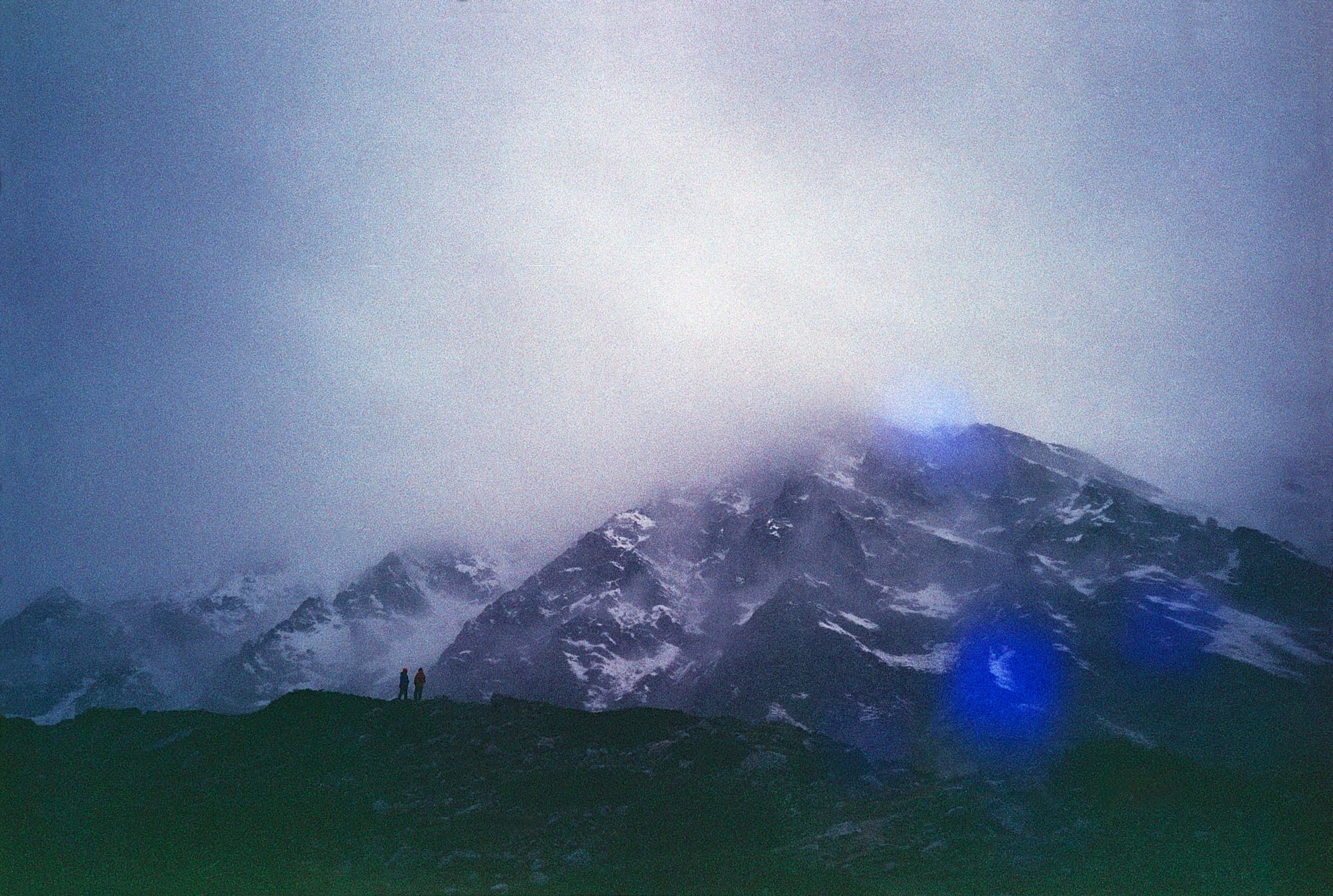 Two people standing on a rocky outcrop at dusk, overlooking a misty mountain range with snow-capped peaks under a dimly lit sky.