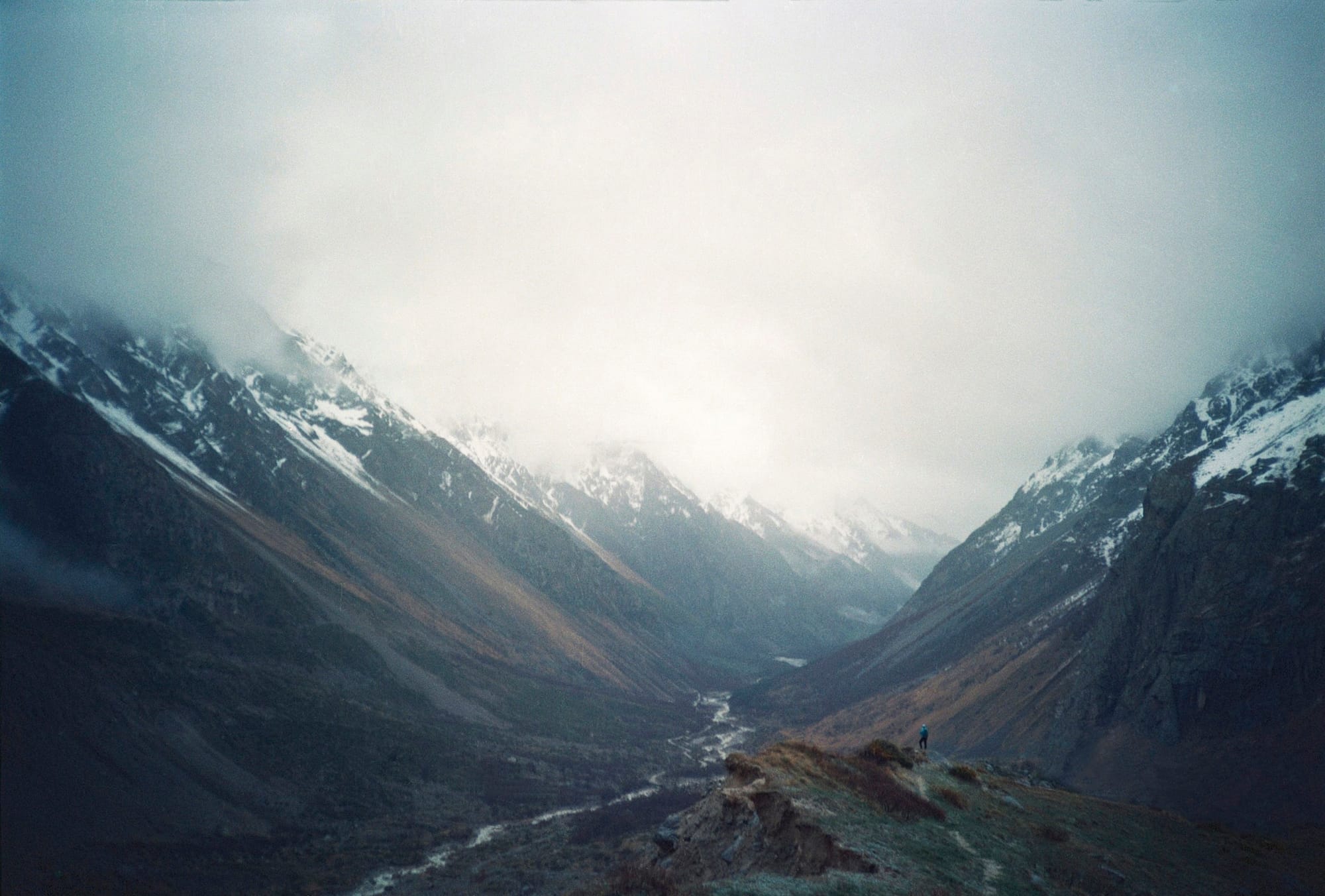 A person standing on a rocky hillside in a mountainous valley, with snow-capped peaks and misty clouds in the background under a cloudy sky.