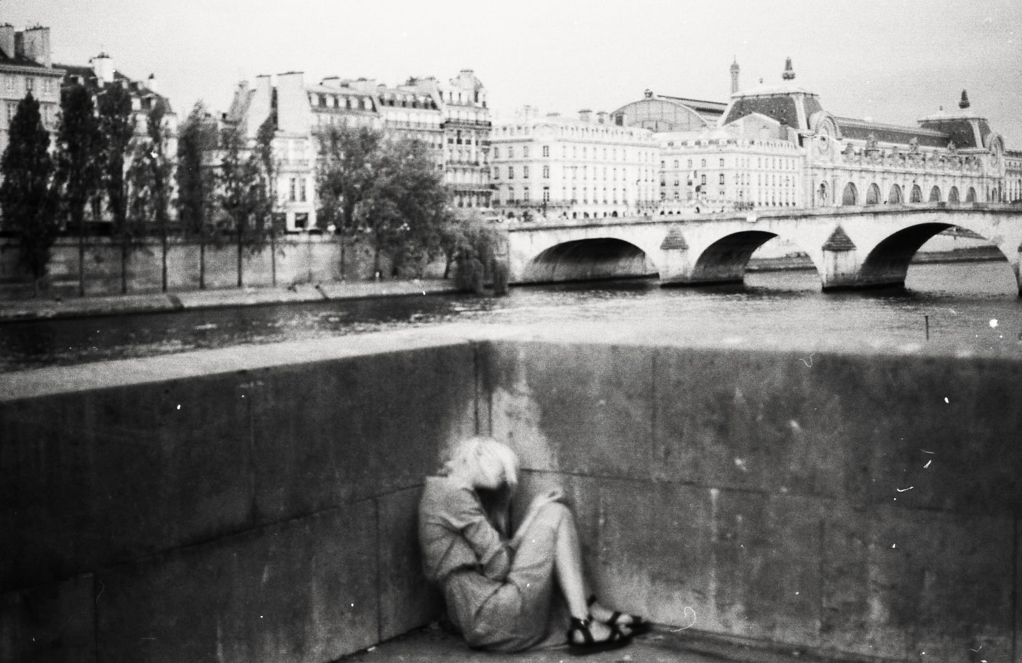 A person sitting alone by a river with a historic bridge and buildings in the background.