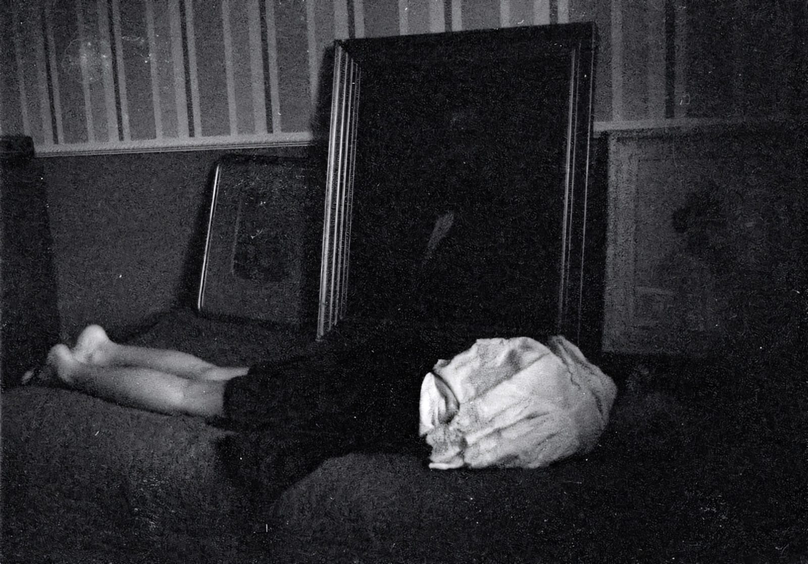 Young woman lying on bed with framed pictures, somber grainy atmosphere.