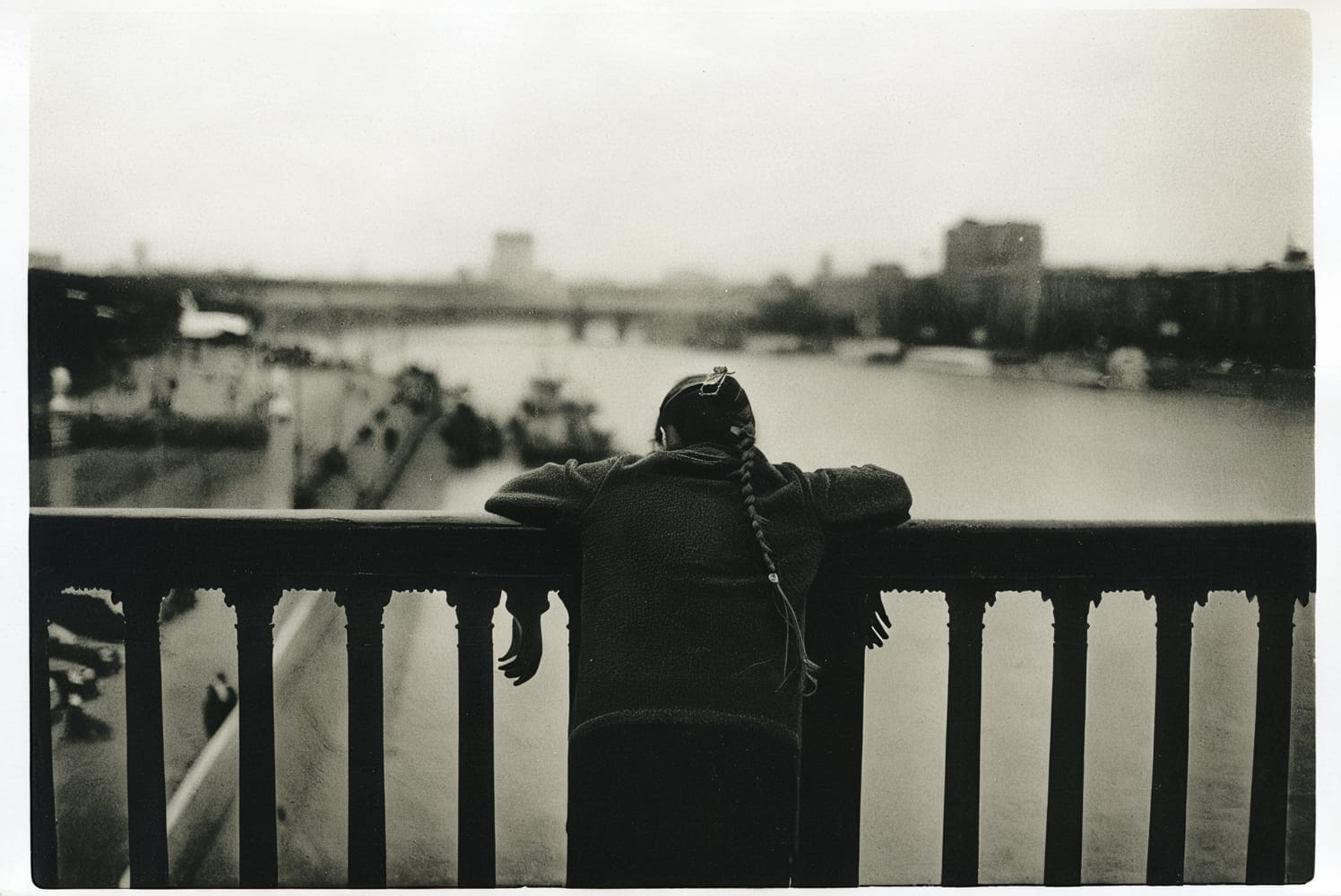 A black and white photograph of a girl leaning on a railing, overlooking a large river.