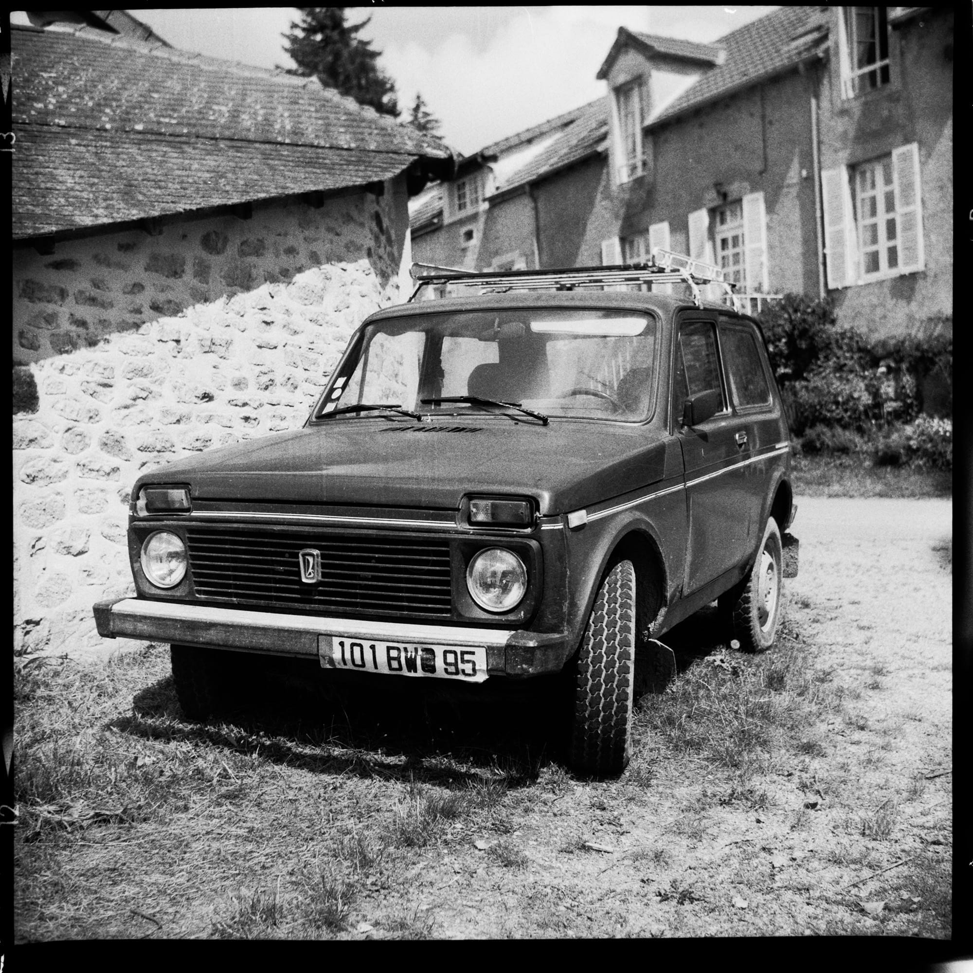 Vintage black and white photo of a classic car parked on a grassy area near old stone buildings.