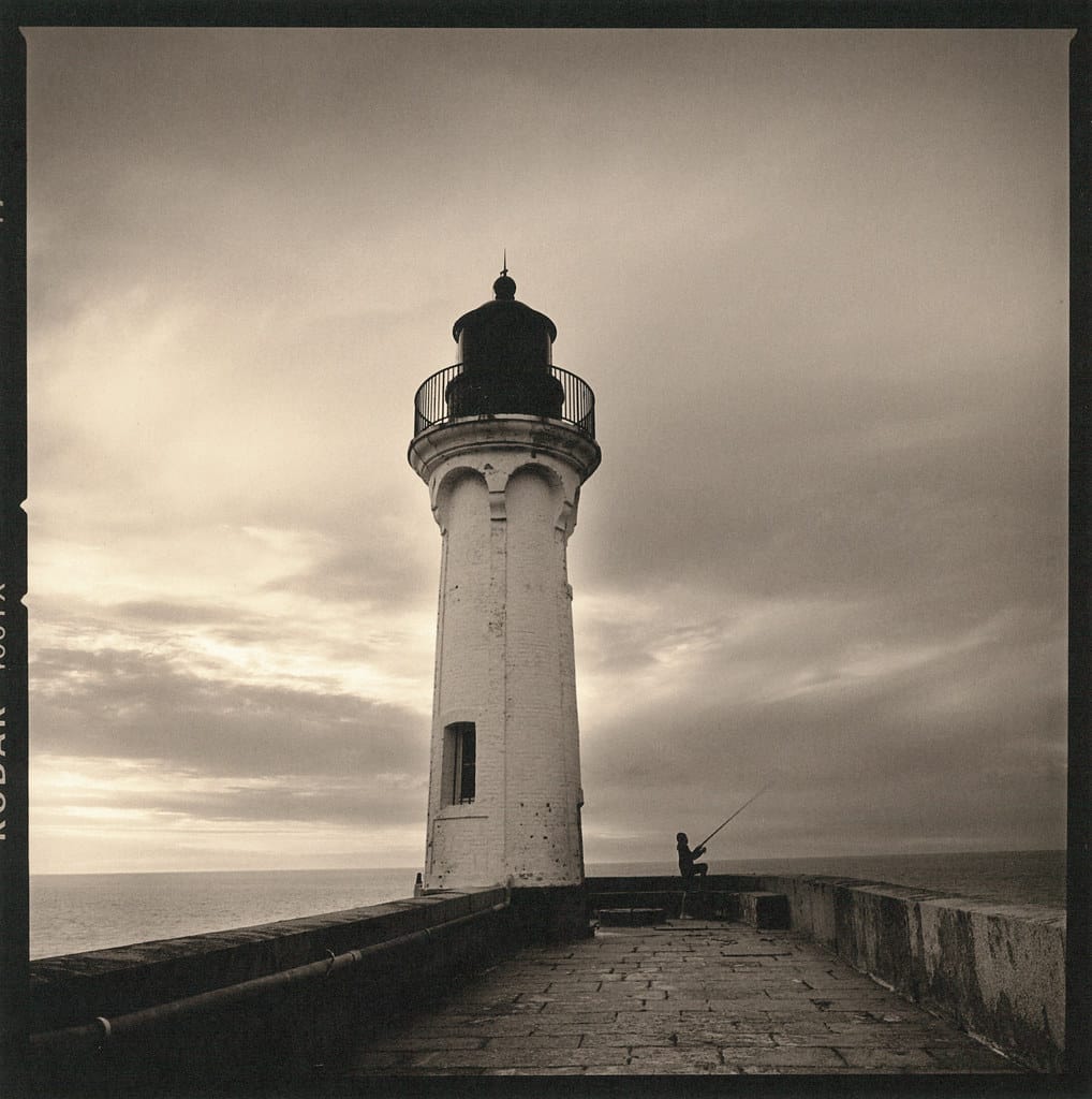 A vintage black and white lighthouse photo with a fisherman on a stone pier.