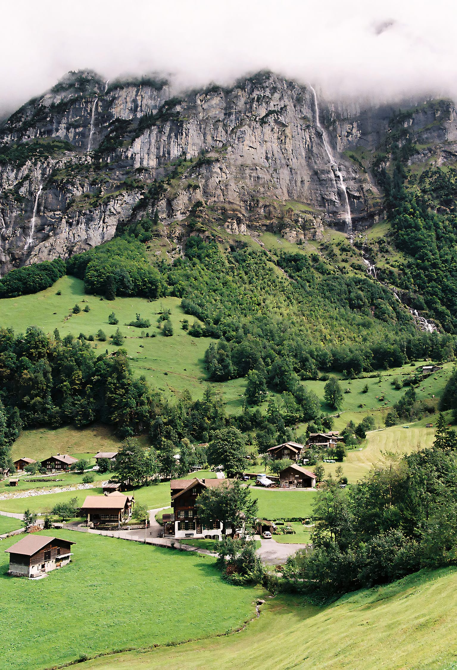 Picturesque village nestled in a lush green valley with towering cliffs and waterfalls in the background.