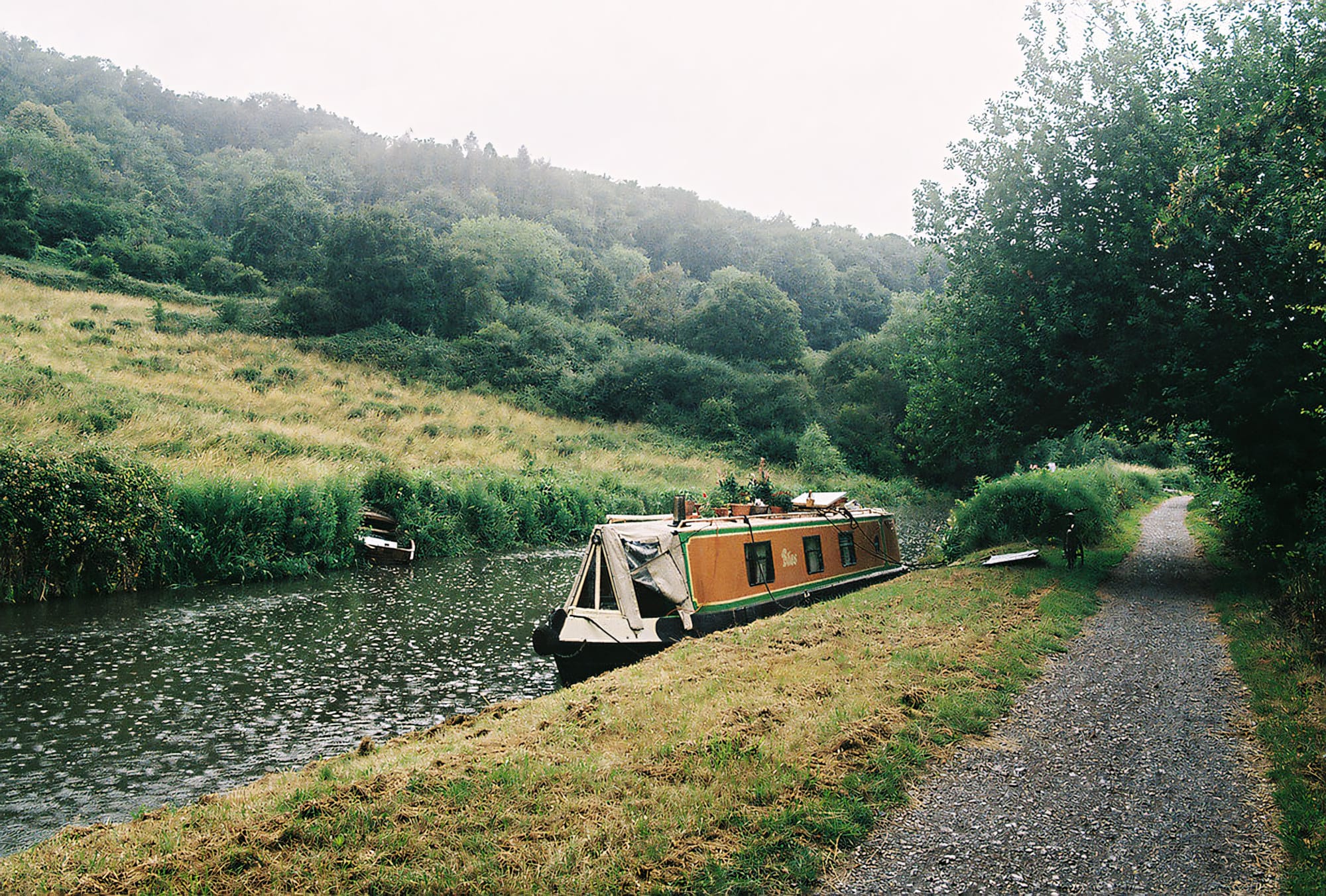 Narrowboat moored on a calm canal, surrounded by lush greenery and a grassy path.