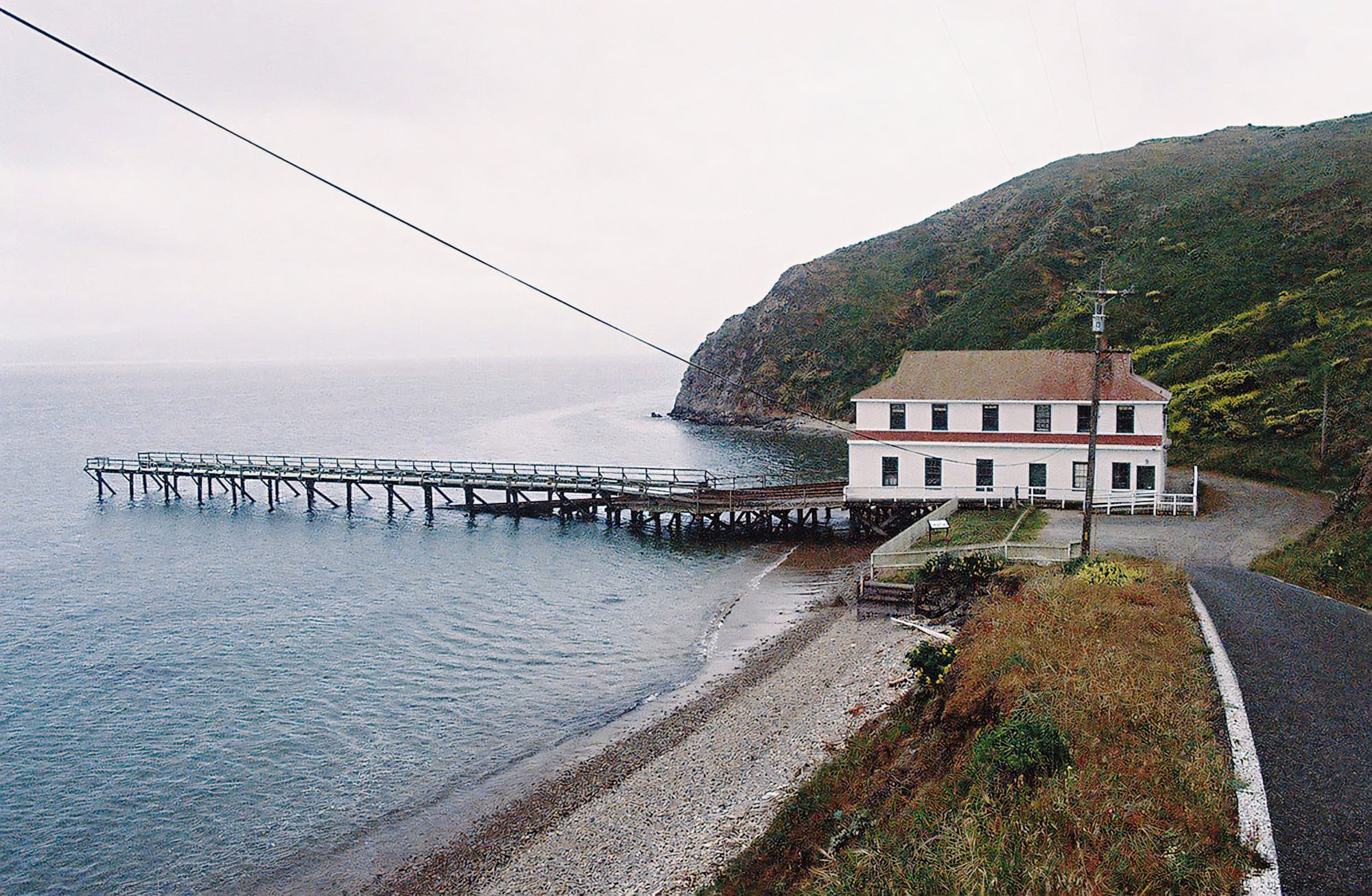 A white building on a pier extending into the ocean, with rocky cliffs and a winding road.