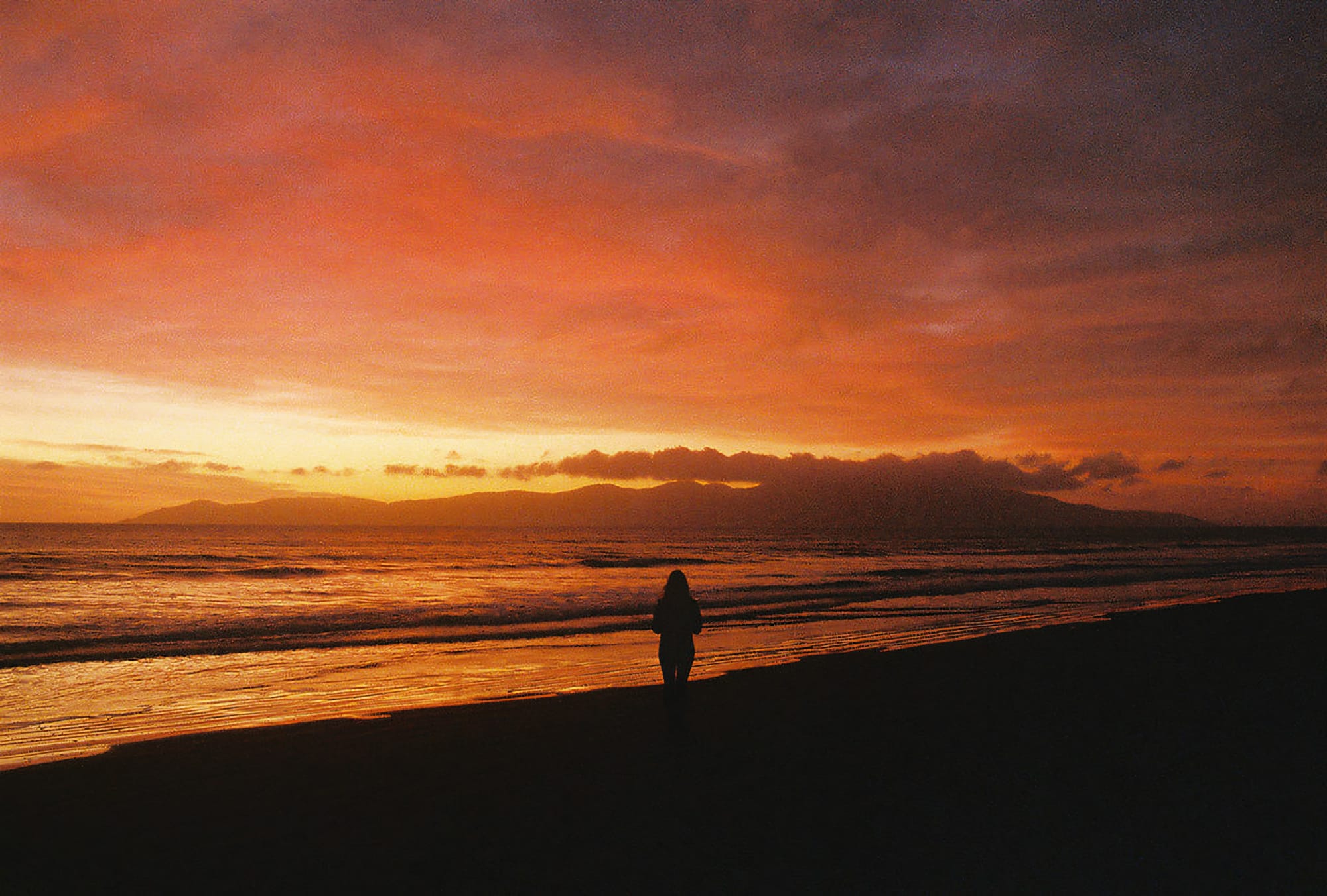 Sunset over the ocean with a person's silhouette on the shore, an island in the distance.
