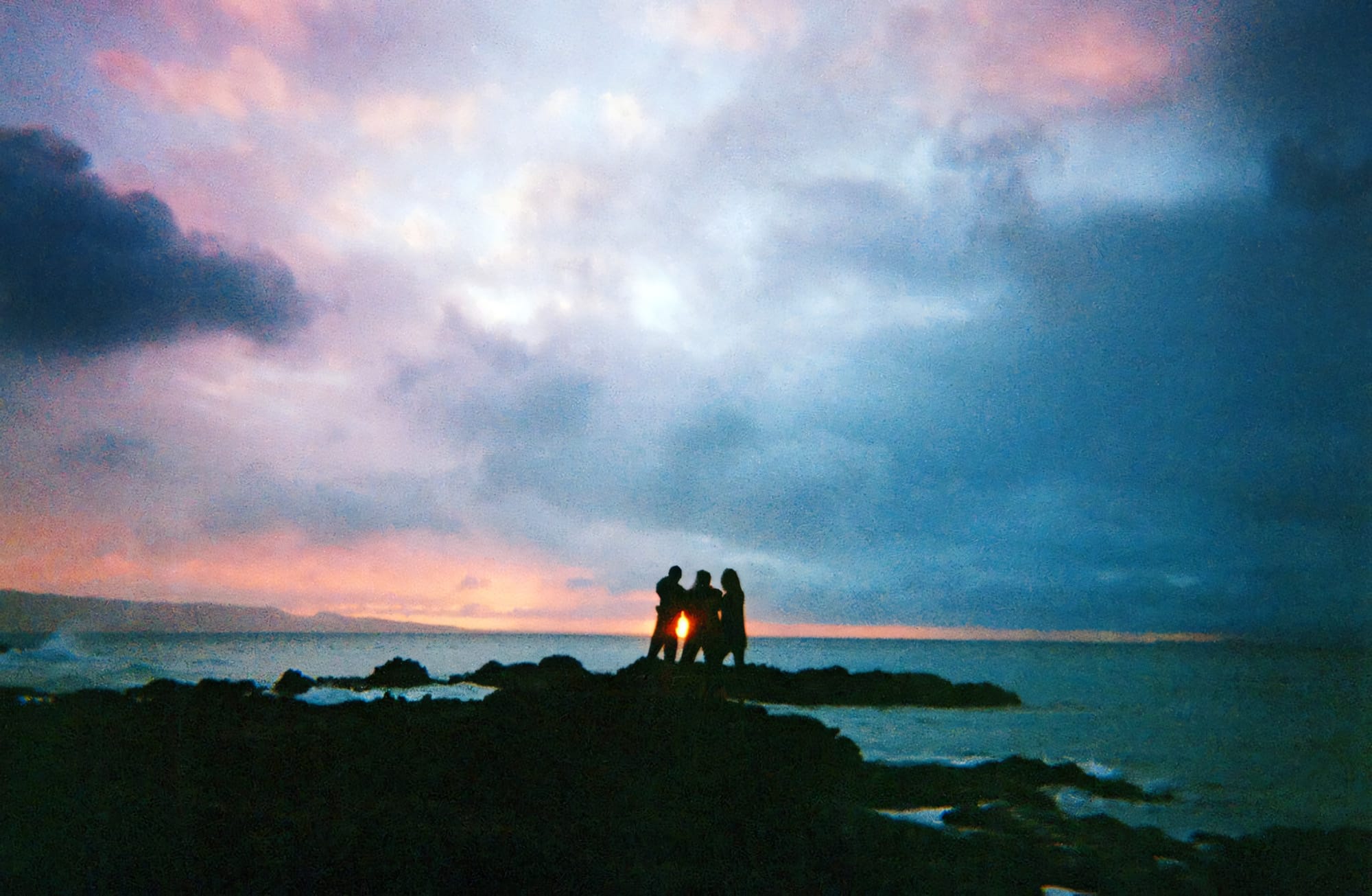 Three figures silhouetted against a vibrant sunset sky, standing on rocks by the sea.