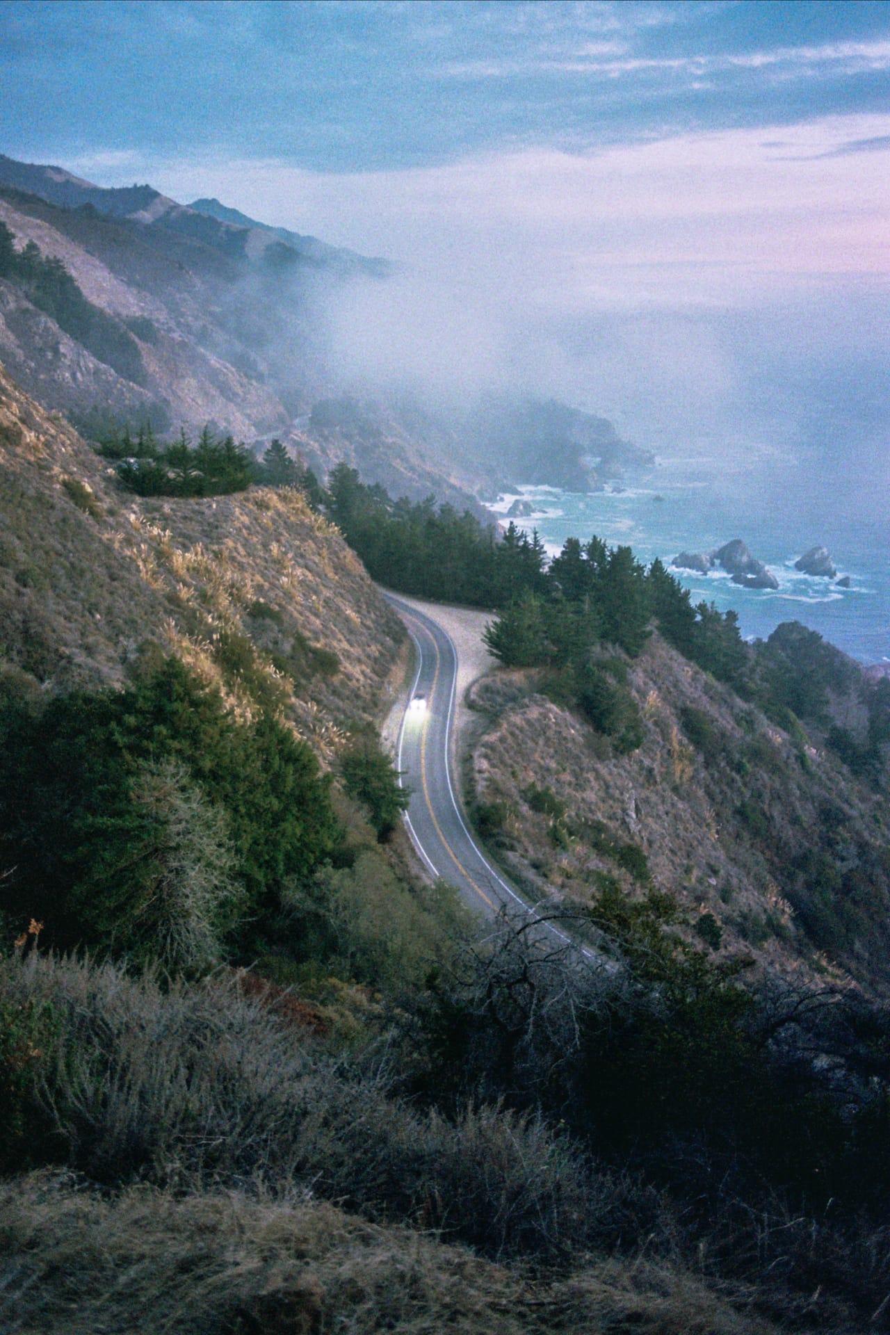 Lone car driving down a winding coastal road with foggy mountains and ocean views.