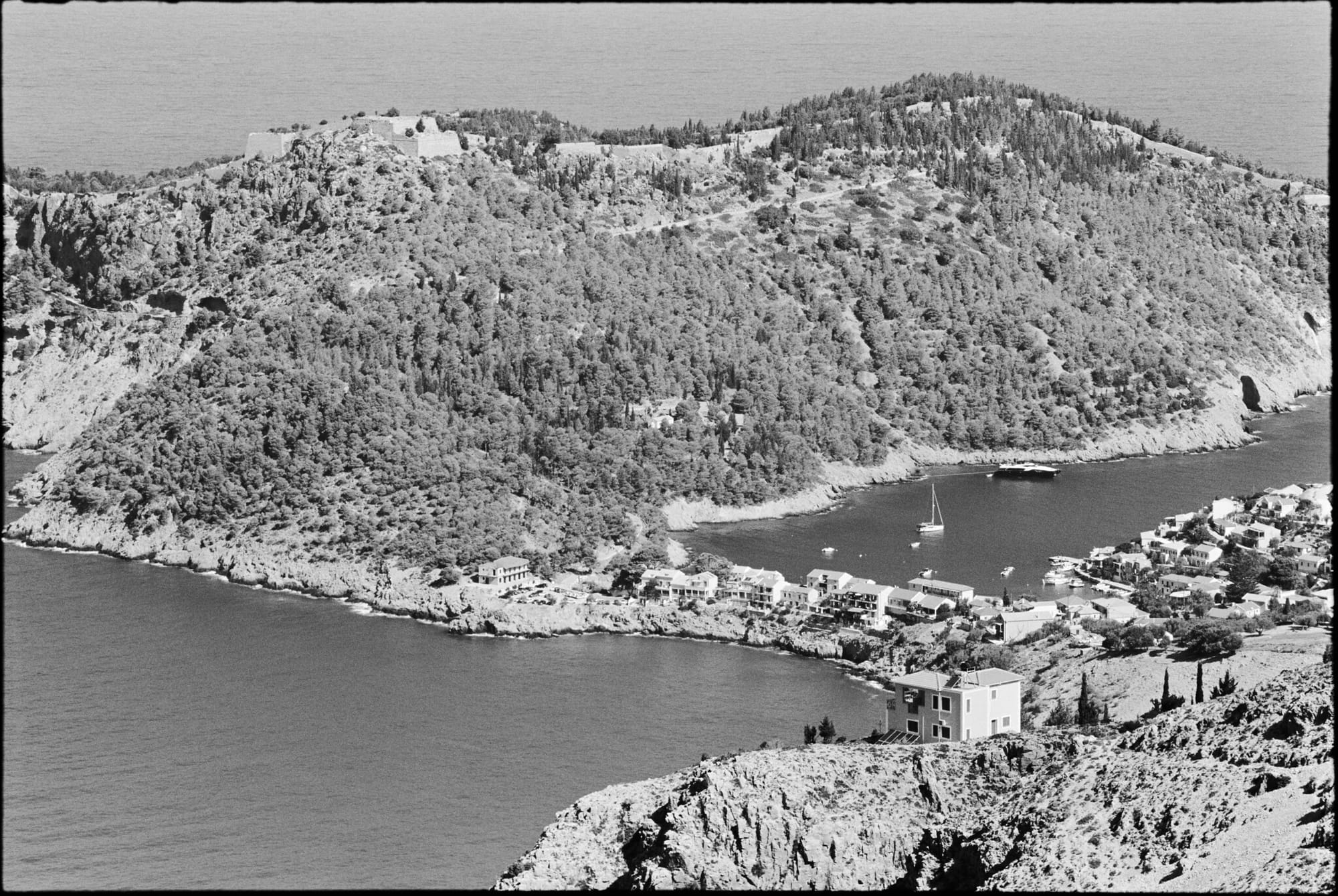 Black and white photo of a coastal village nestled at the base of a forested hill.