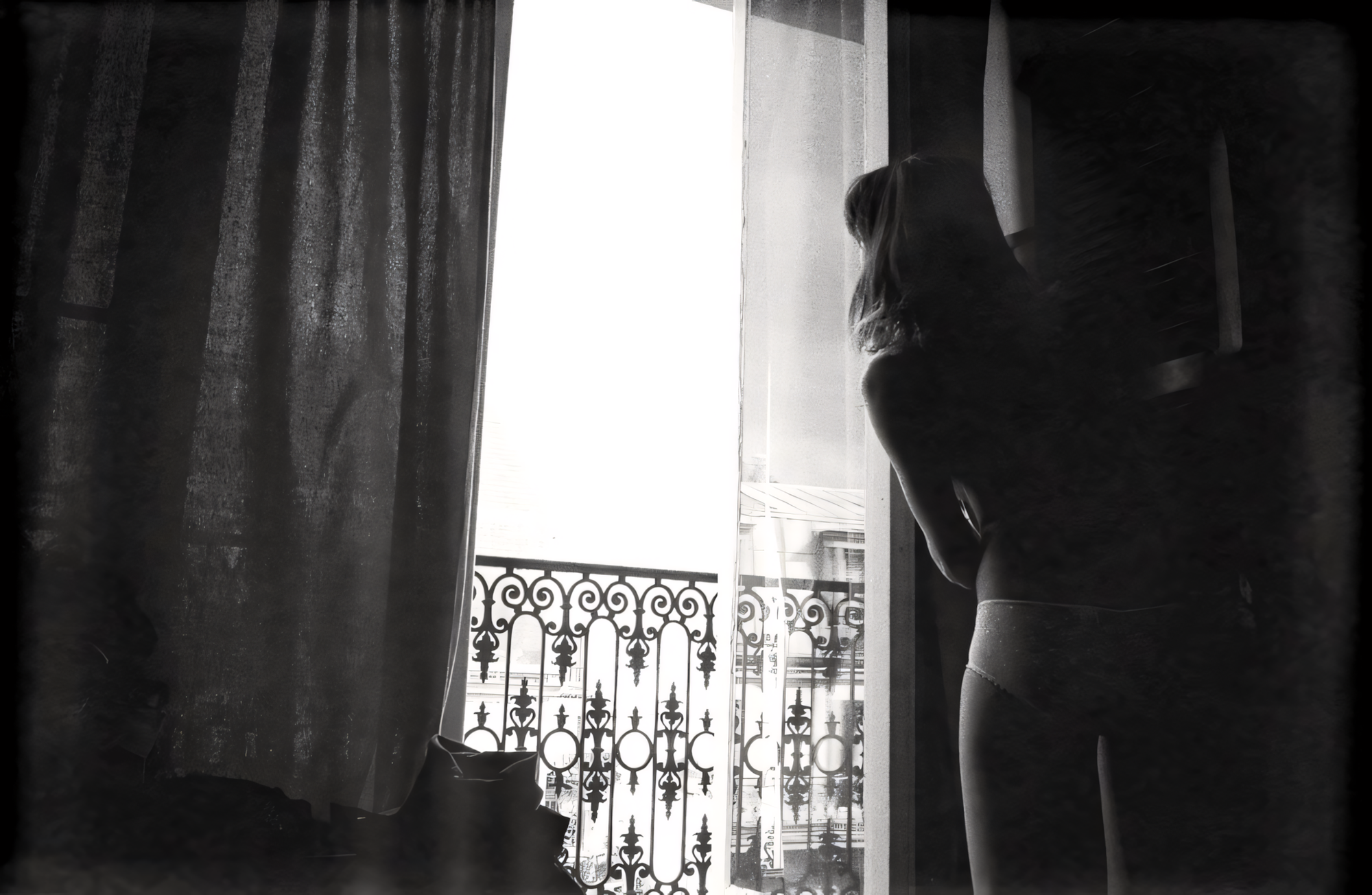 Woman in underwear standing by a window with ornate wrought iron balcony.