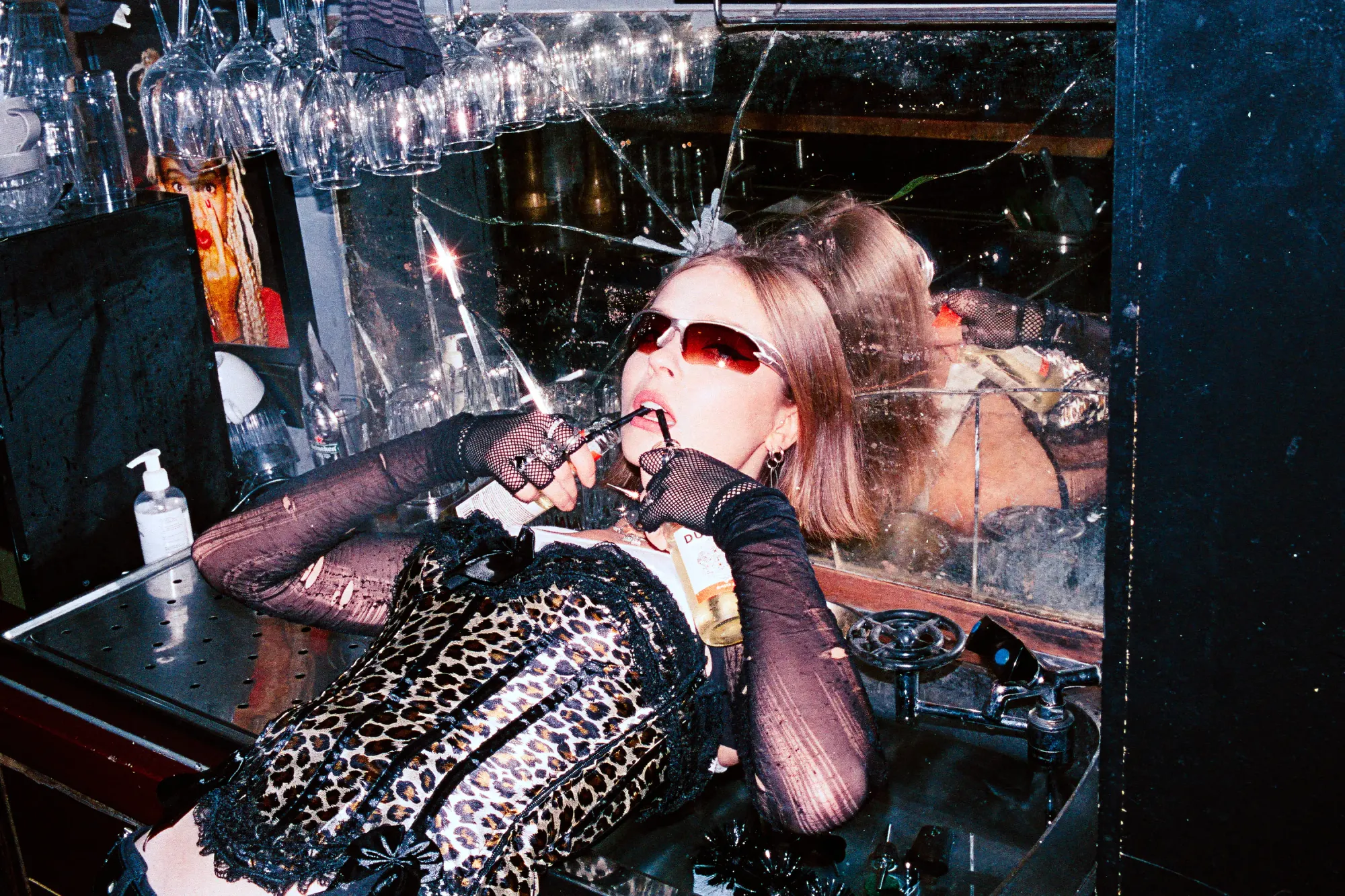 Young woman in a leopard print outfit and sunglasses in a reflective bar setting.