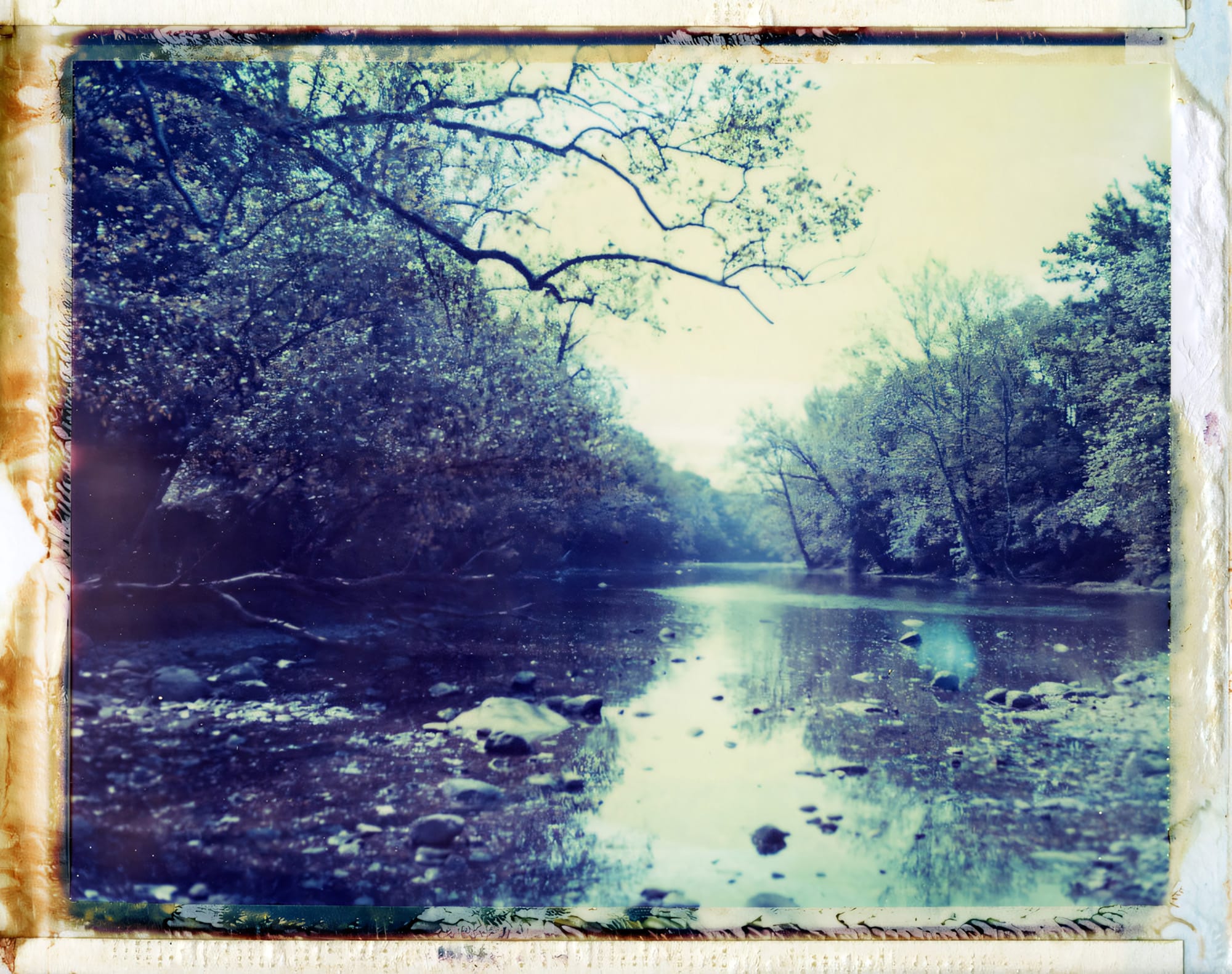 Rocky stream reflecting trees with cyan tones on Polaroid.