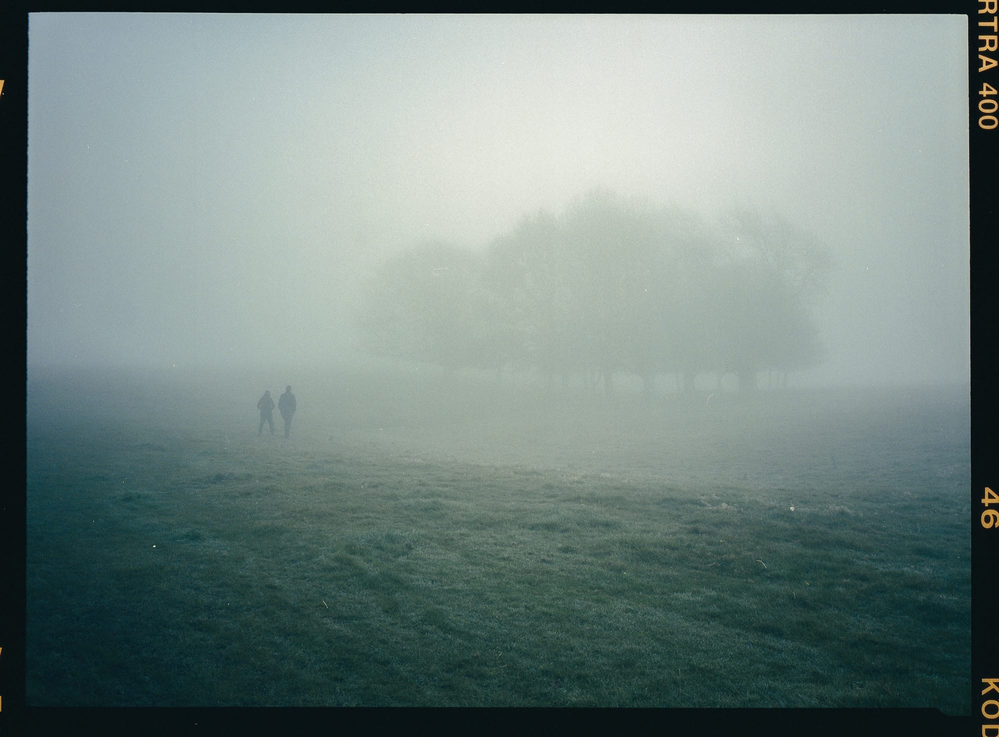Two figures walking across foggy field toward distant trees on film.