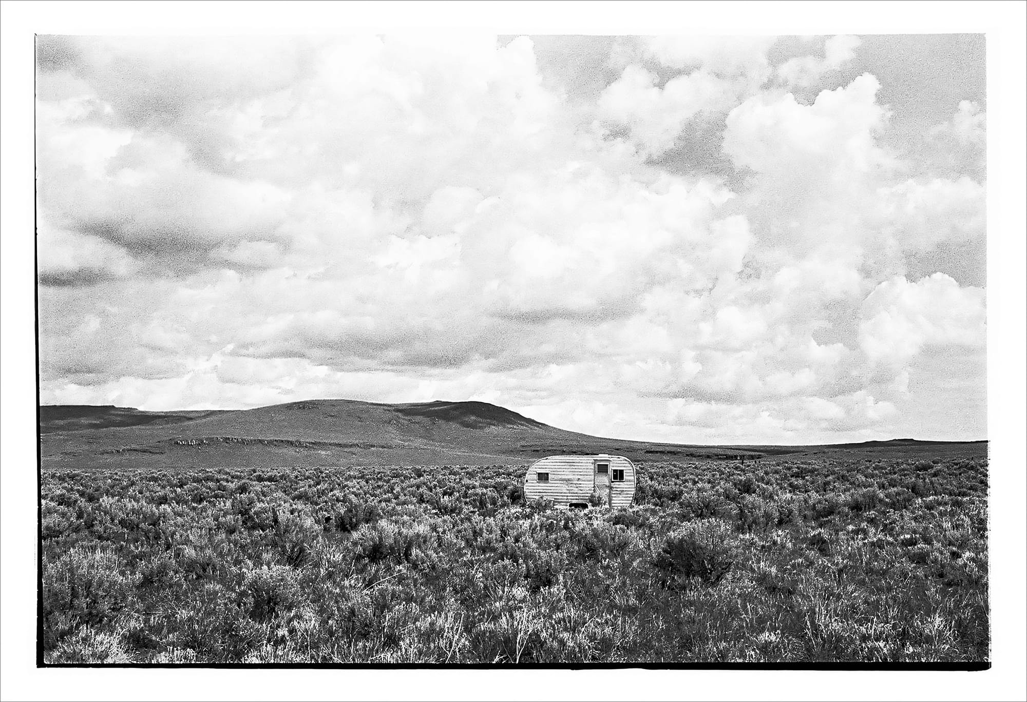Black and white photo of a small, weathered trailer in a vast, open field.
