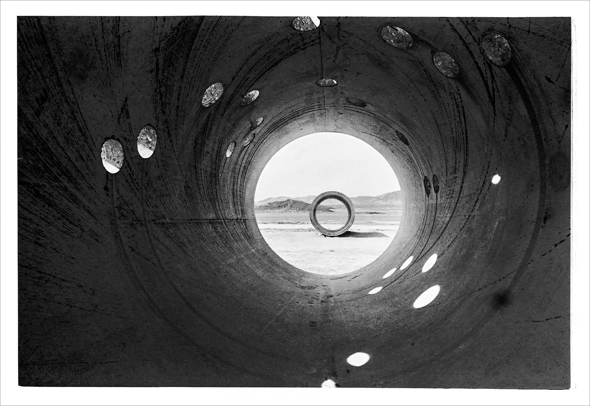 Monochrome image of a cylindrical tunnel leading to a barren, mountainous terrain.