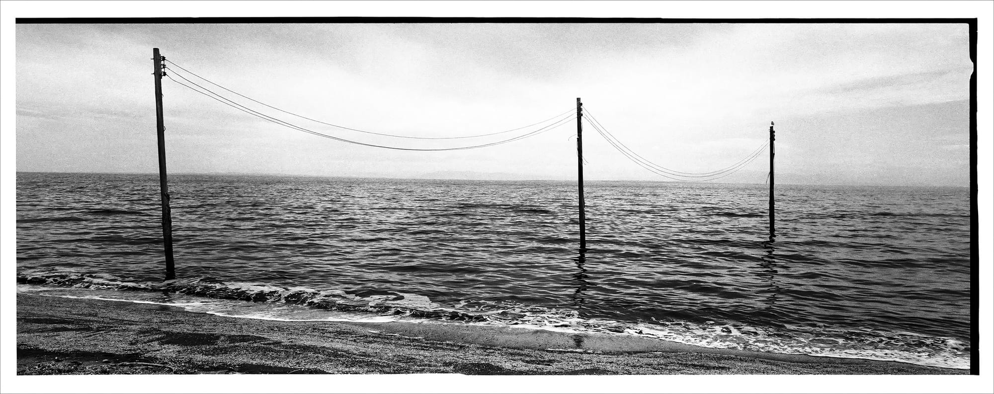 Vintage scene of power lines with poles in water near the coastline.