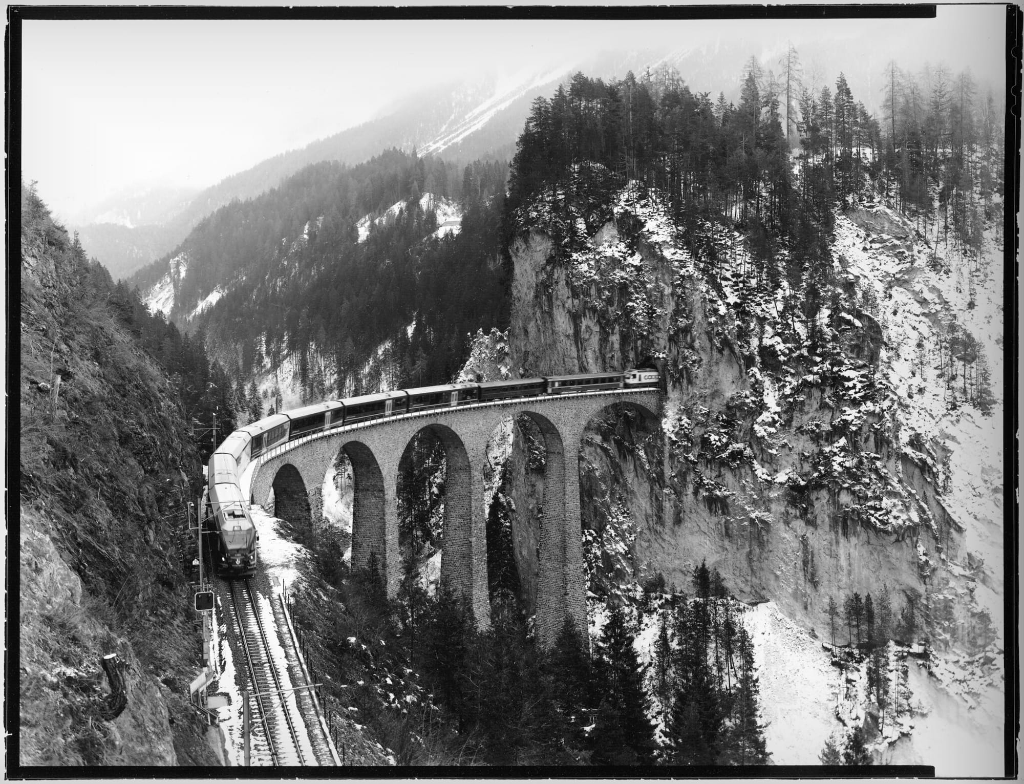 Train crossing stone viaduct through snowy mountain valley.