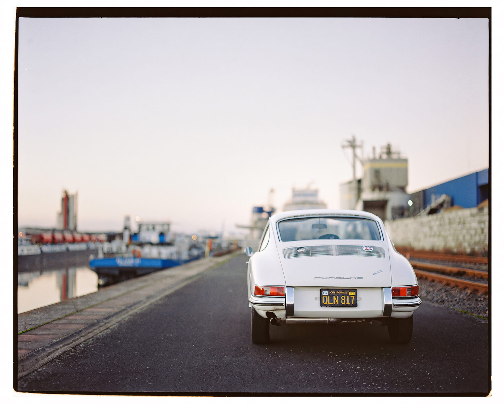 A classic white Porsche by a waterfront, with boats and industrial buildings visible.